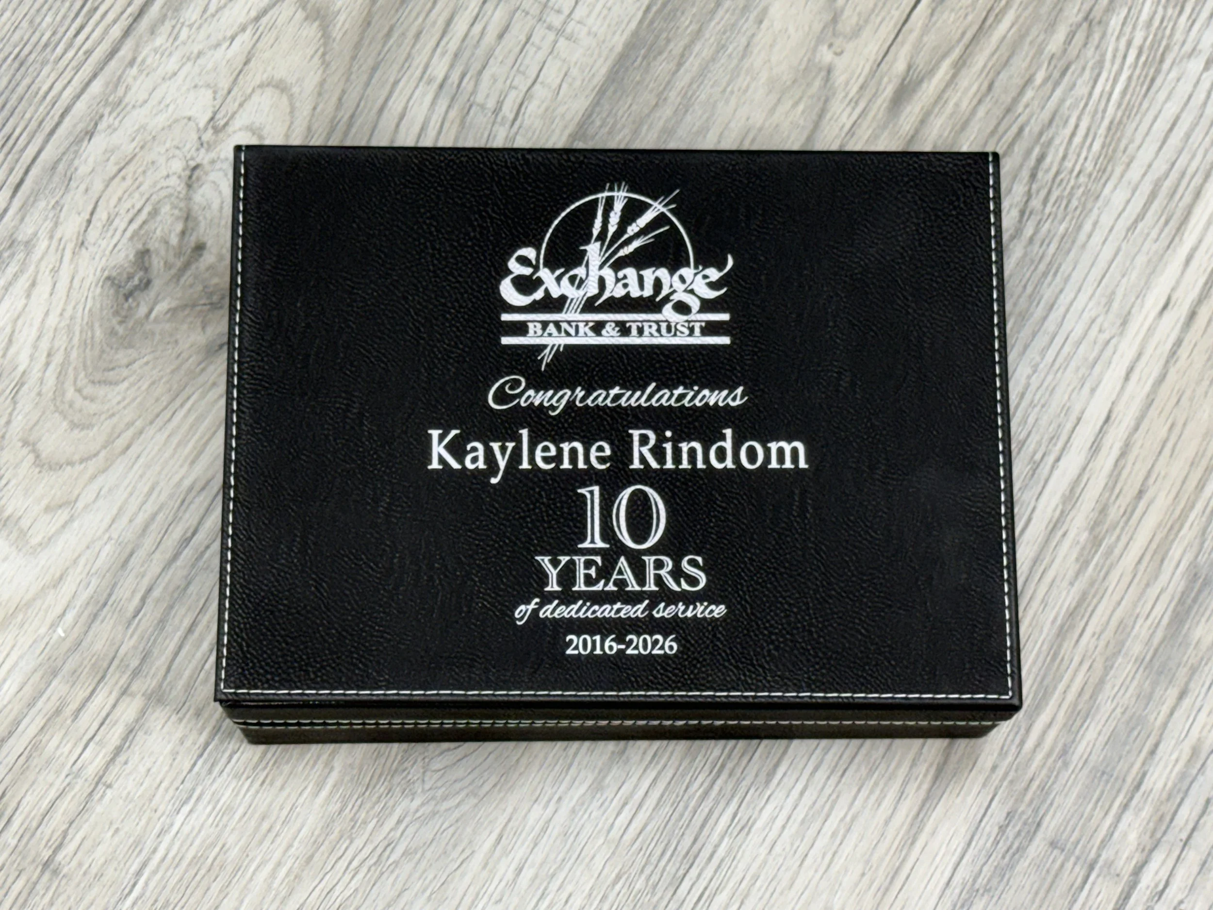 Black plaque or certificate with white text and logo, placed on a light wooden surface. It congratulates Kaylene Rindom on 10 years of dedicated service from 2016 to 2026.