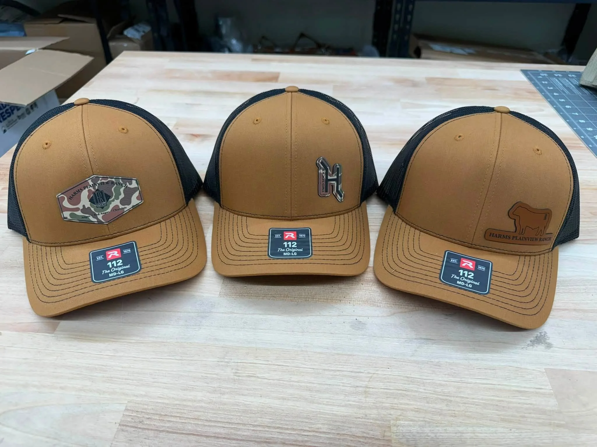 Three brown and black trucker hats on a wooden table, with labels indicating they are original 112 models from Harms Plainview Ranch.