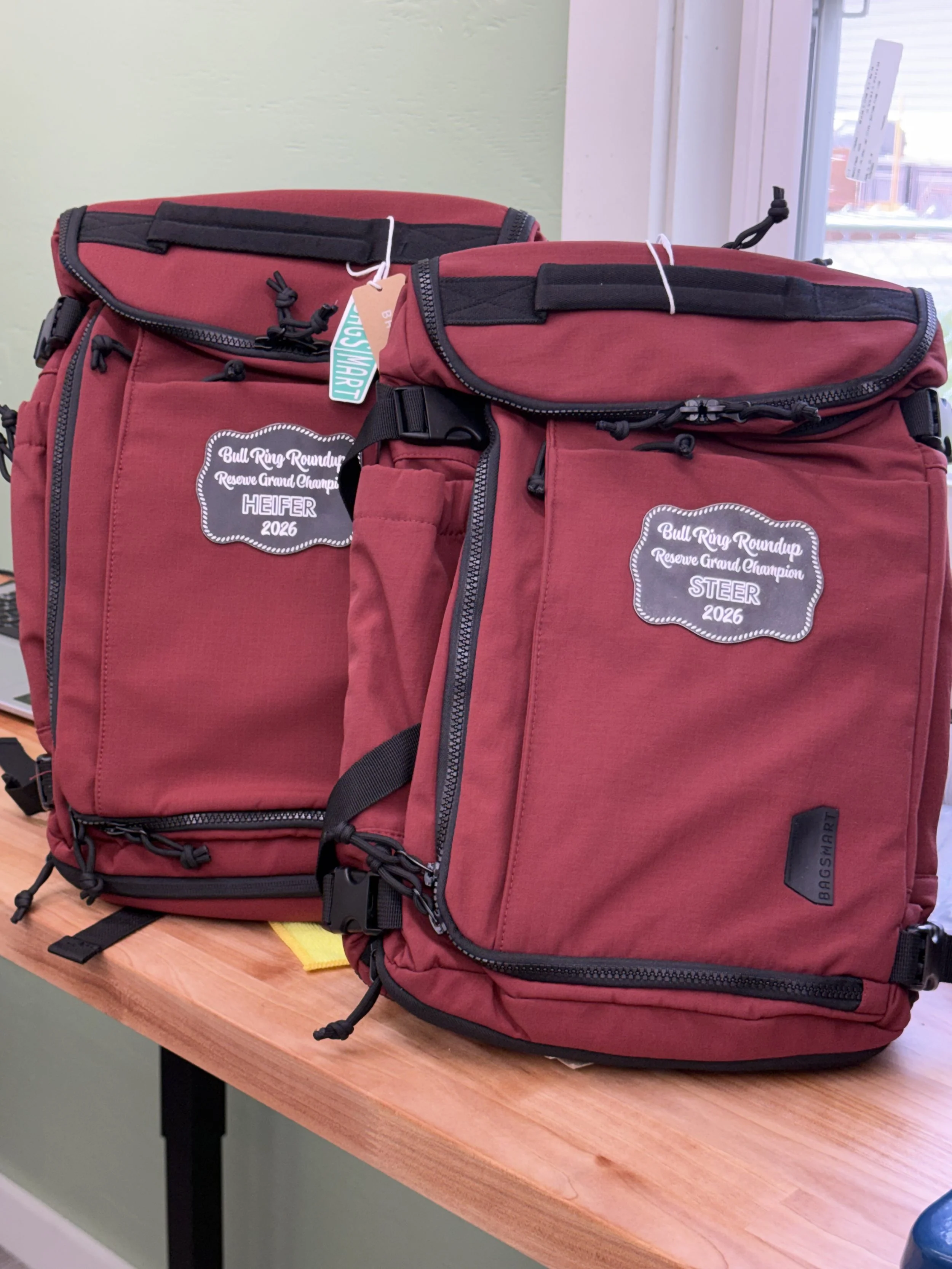 Two red backpacks with black zippers and straps on a wooden table, with a window in the background.