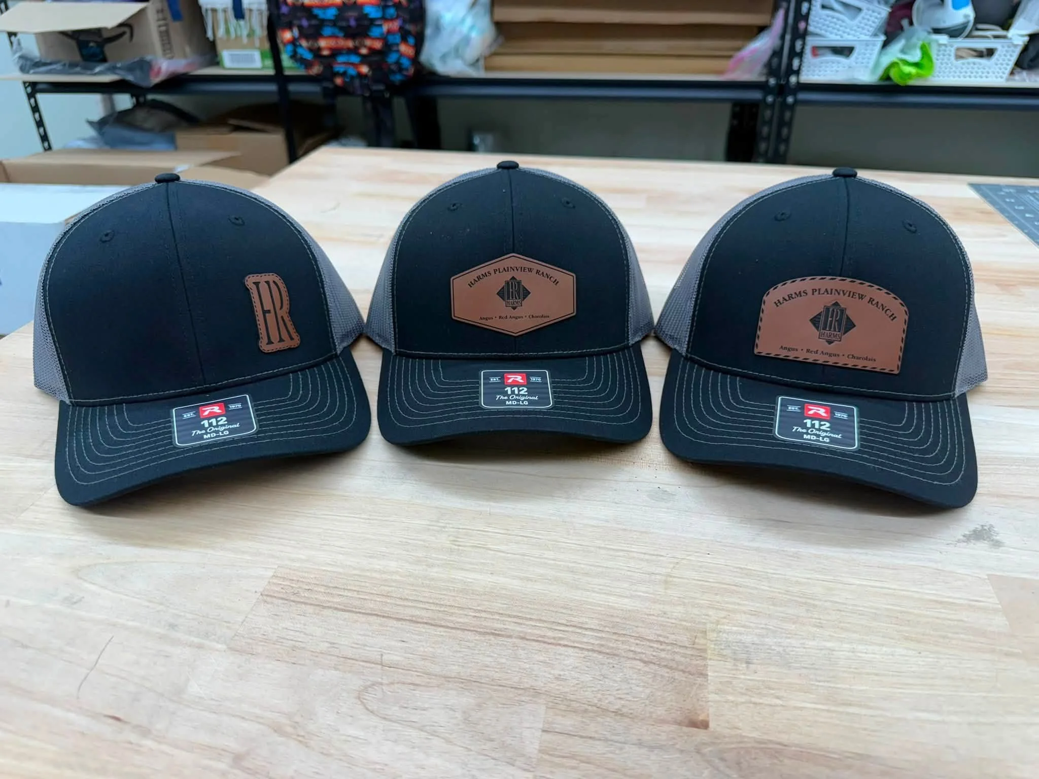 Three black baseball caps with tan leather patches on the front, placed on a wooden surface in a store or warehouse setting.