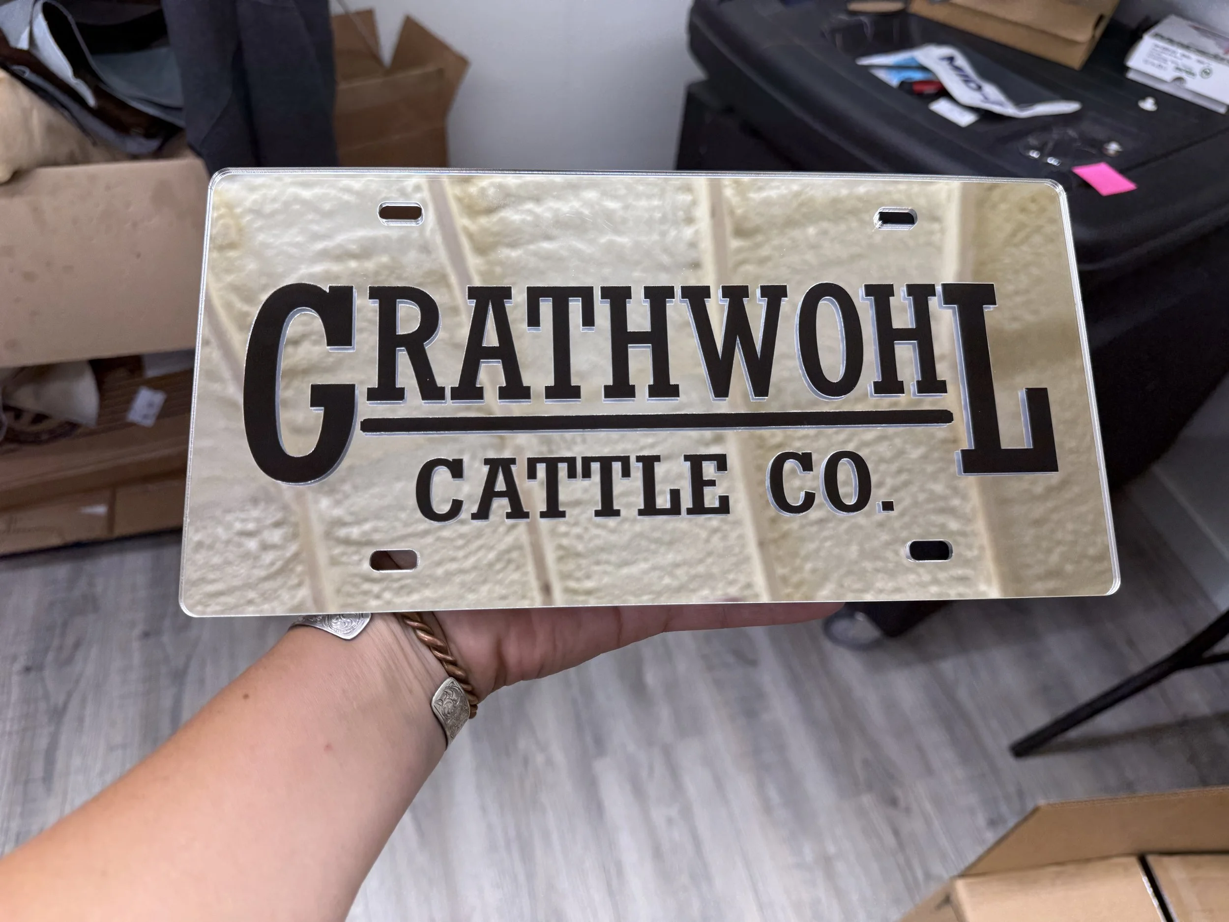 A person holding a metal license plate frame in front of a cluttered background with boxes and office supplies. The frame has cutouts for a license plate and features black text reading 'CATAHUEY, CATTLE CO.'