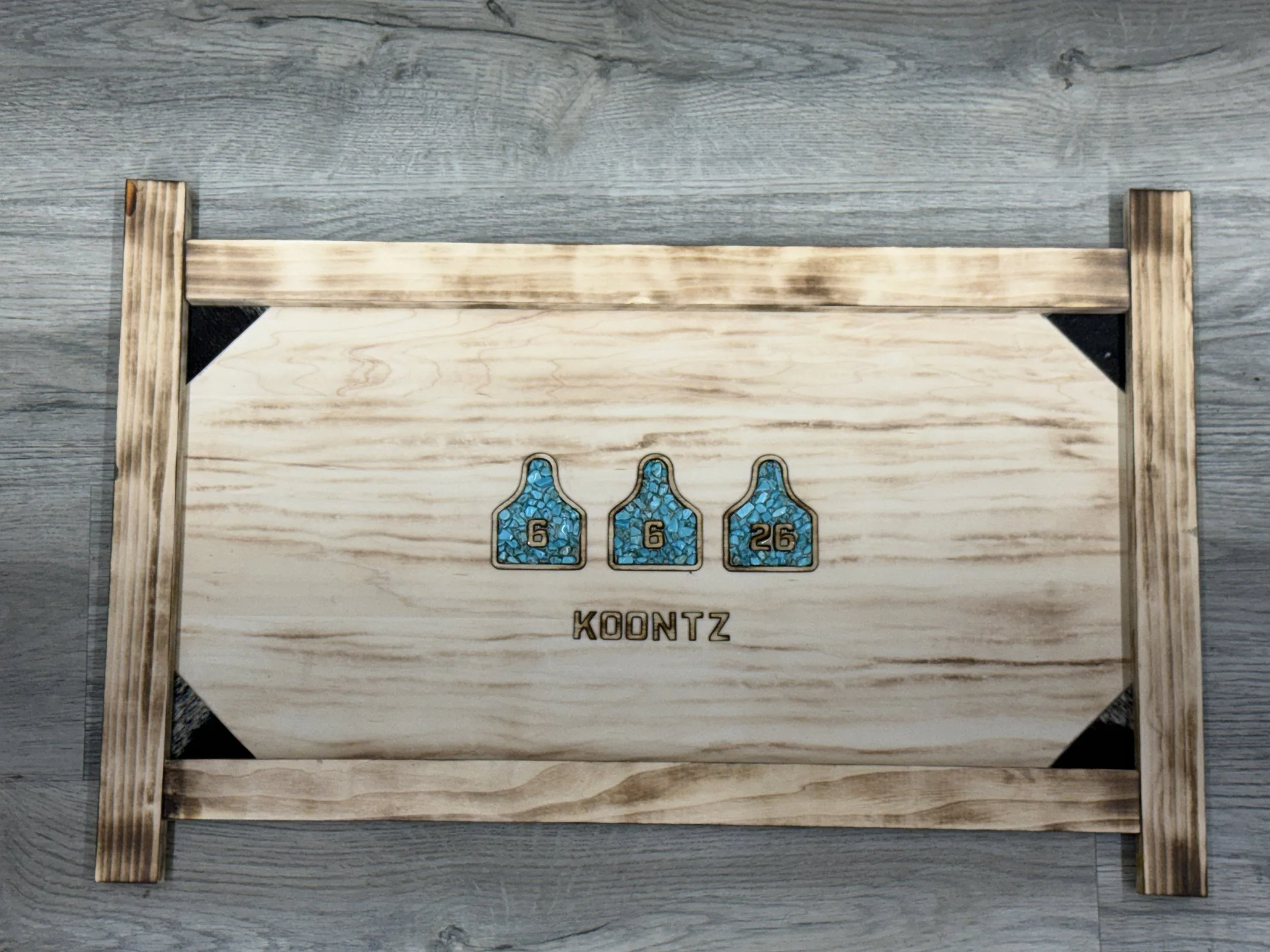 A wooden game board with blue mosaic tiles and numbers 6, 6, and 26 on it, supported by four wooden legs and placed on a gray wooden surface.