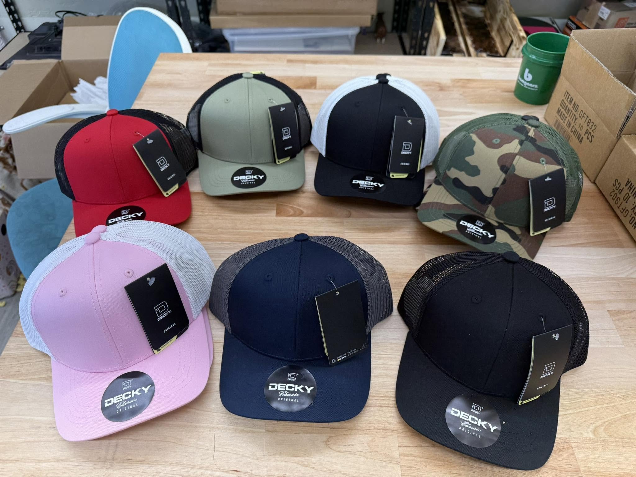 Display of various baseball caps in pink, navy, black, camouflage, and gray colors on a wooden table.