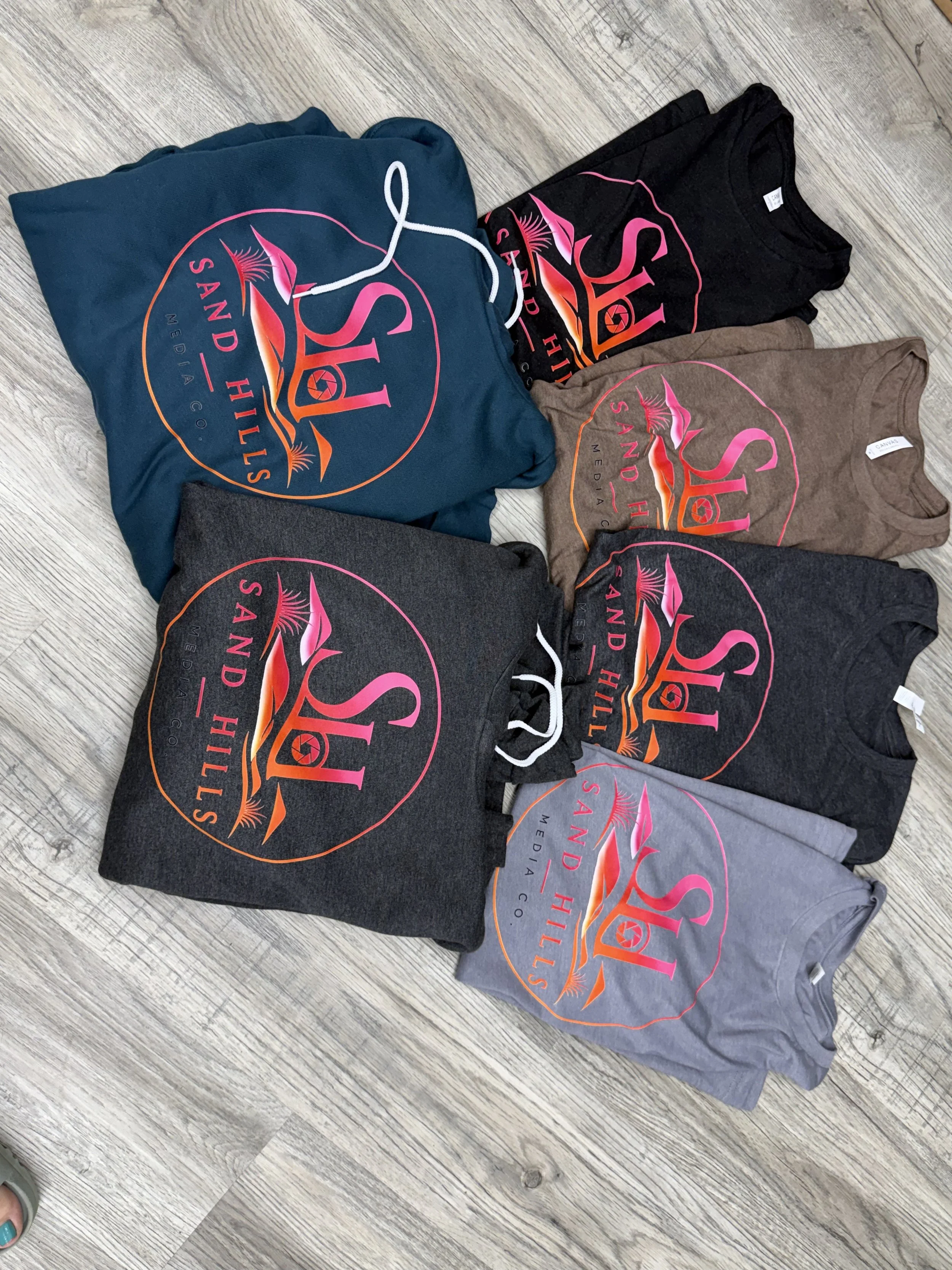 Multiple folded t-shirts in various colors with a large, colorful logo on the front, laid out on a wooden surface.