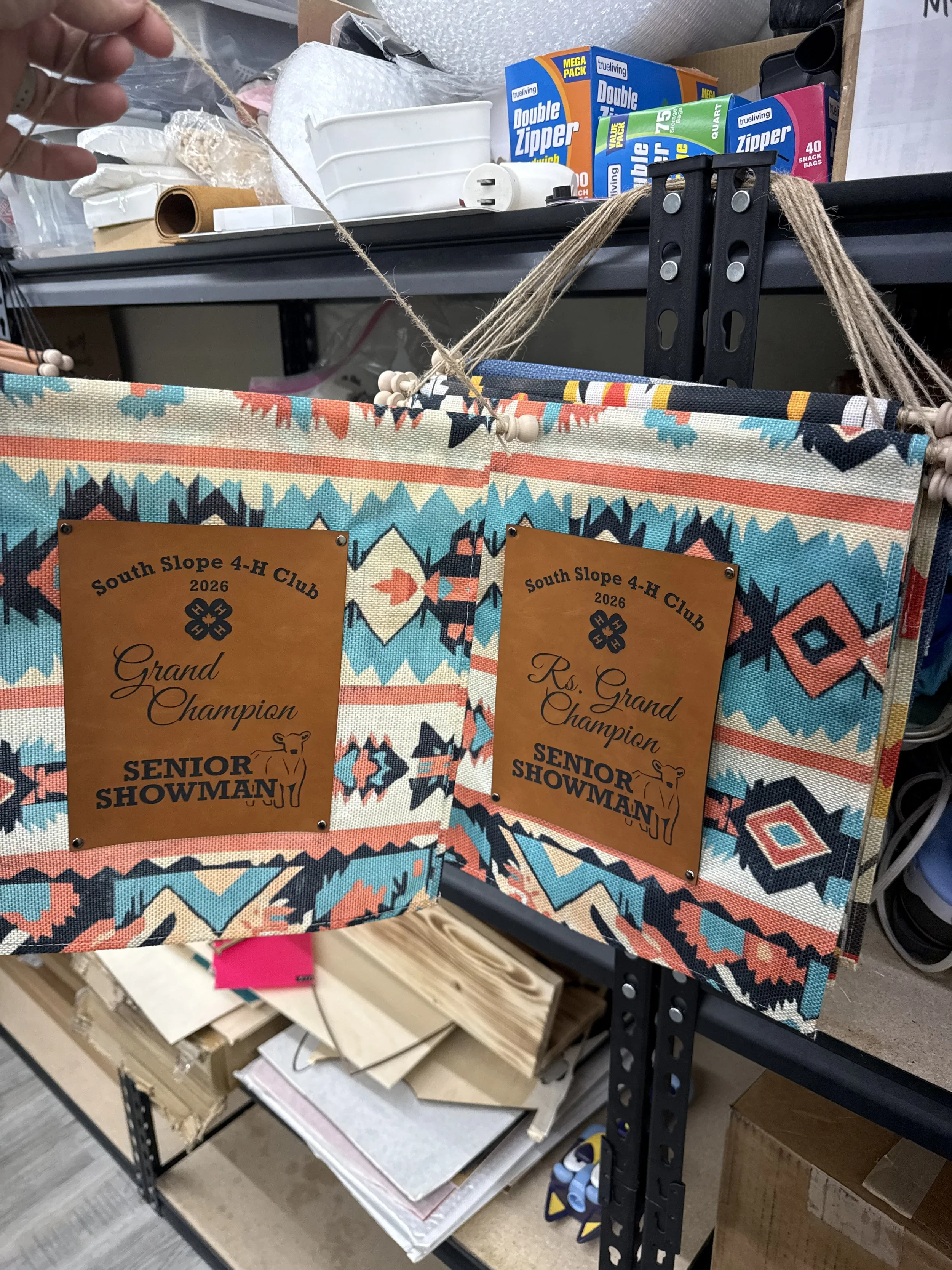 A workbench with a fabric basket displaying two orange certificates labeled "South Slope 4-H Club 2026 Senior Swimman". The basket is decorated with colorful, southwestern-style patterns. The background includes boxes of Zipper storage bags and some other miscellaneous supplies.