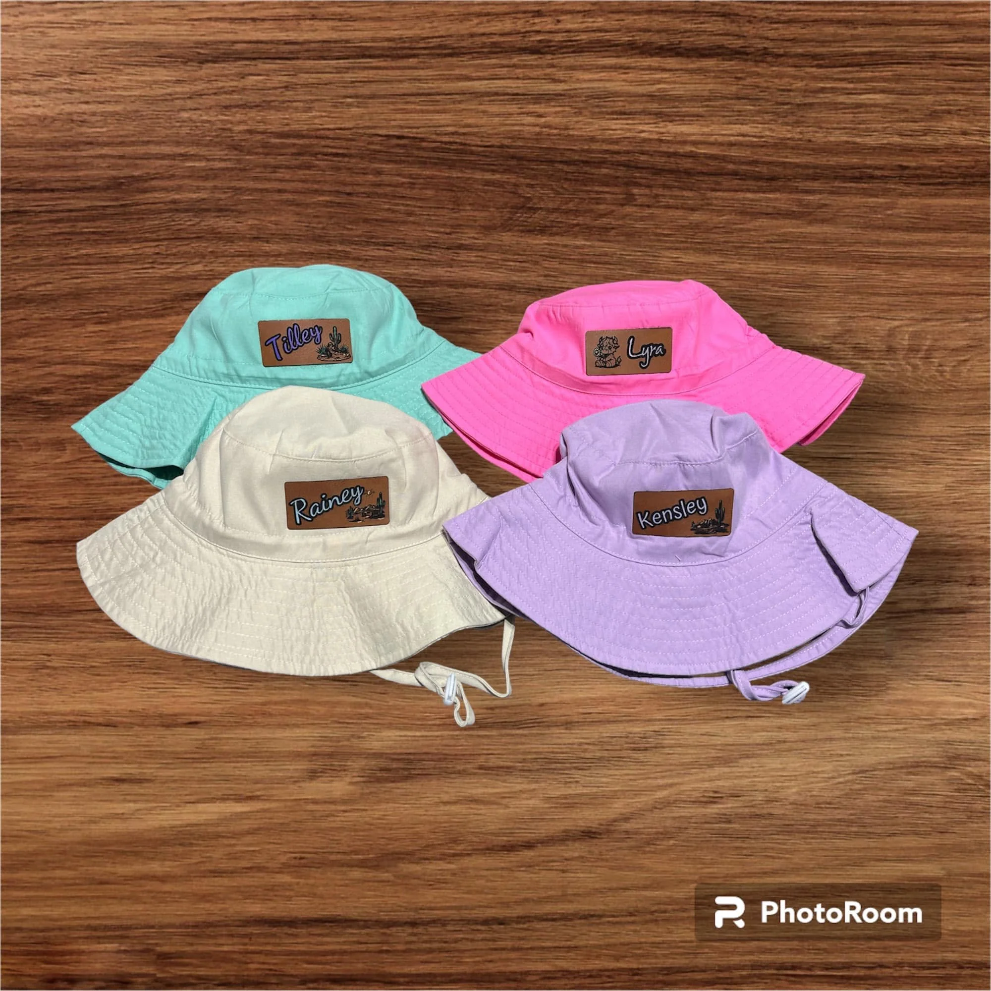 Several pastel-colored bucket hats with stitched brims and labels with names and small cactus or lion illustrations, laid out on a wooden surface.