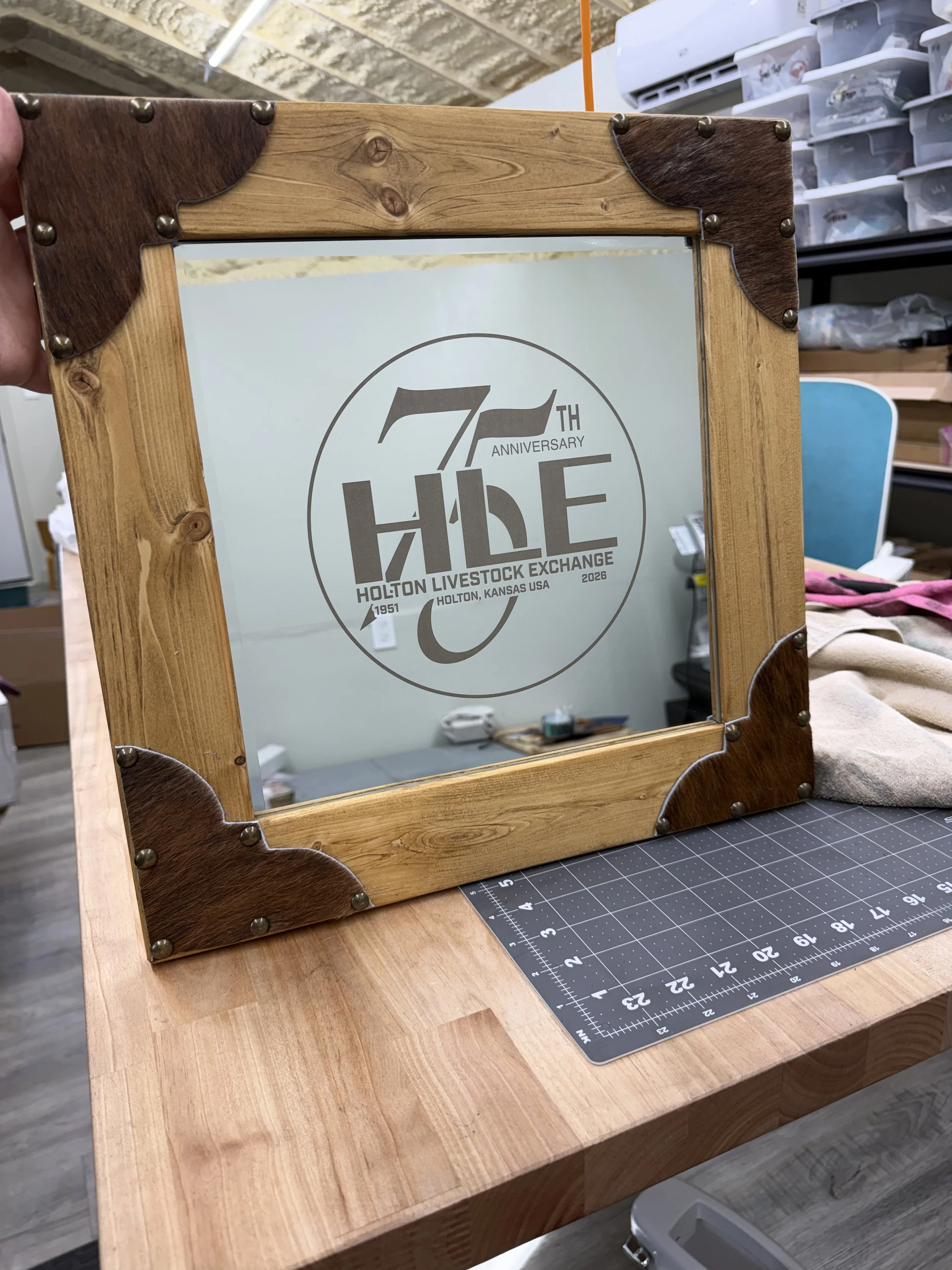 A wooden tabletop featuring a decorative mirror with a wooden frame, engraved with a logo commemorating the 70th anniversary of the Holton Livestock Exchange, located in Holton, Kansas, in 2026. The mirror's frame has dark wood corner accents secured with metal fasteners, and a cutting mat with measurement markings is visible underneath.
