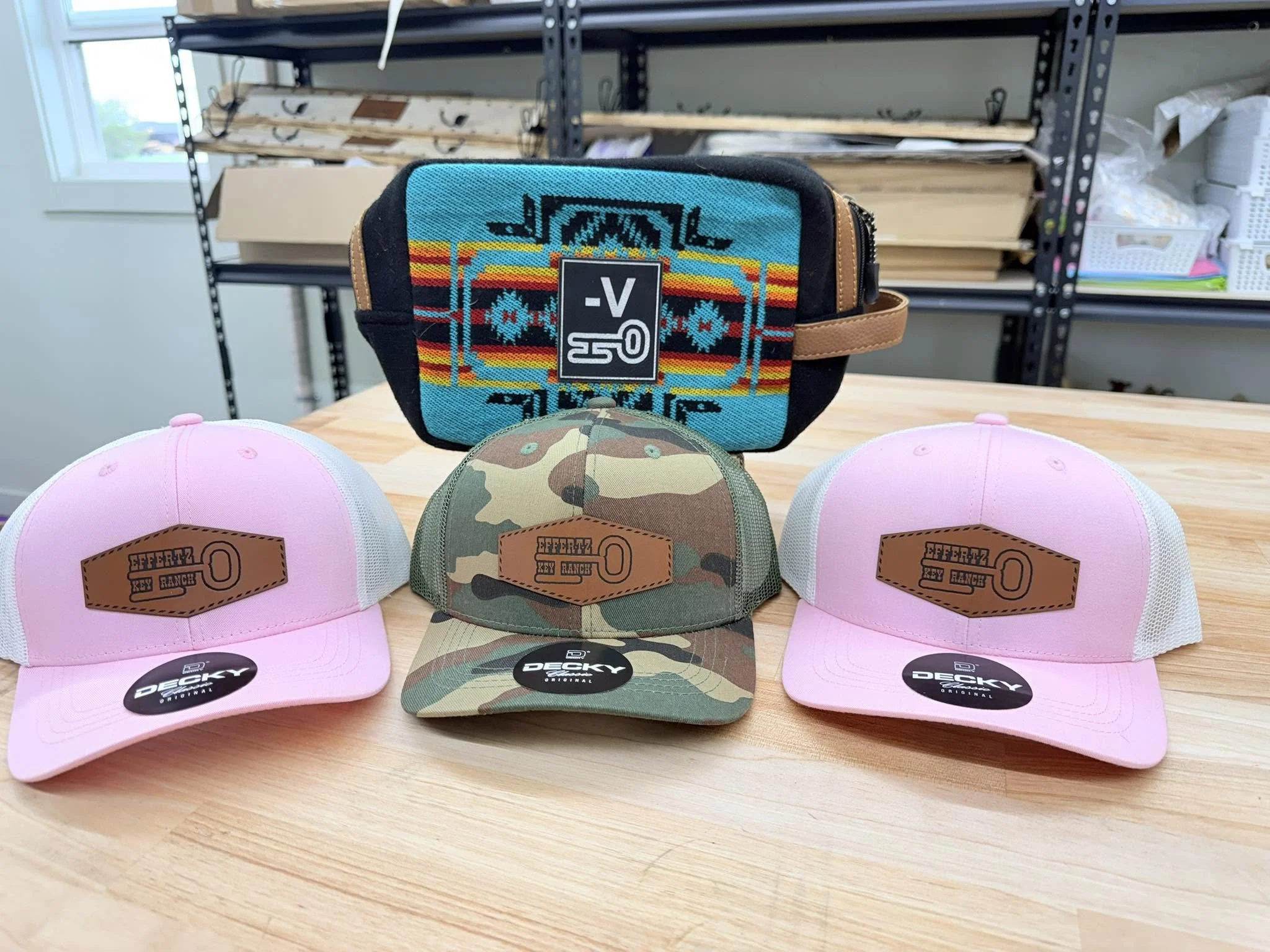Four caps on a wooden table: two pink and two camouflage, with a black cap behind, opposing the pink caps.