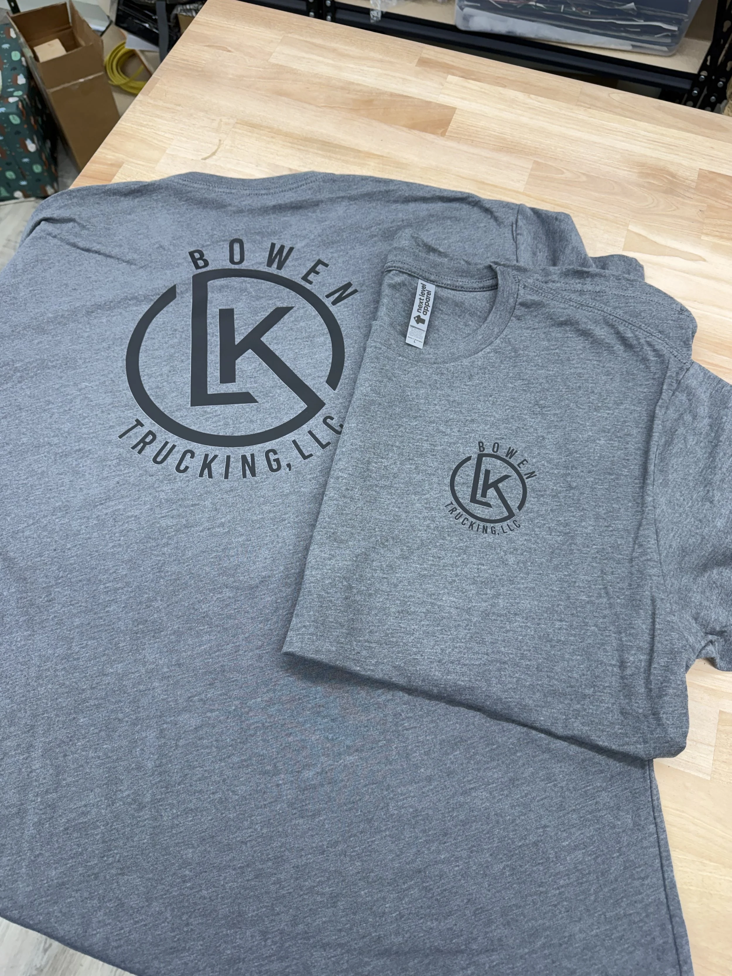 Gray t-shirt with a black circular logo that reads 'BOWEN TRUCKING, LLC' and a large 'LK' in the center, lying on a wooden surface.