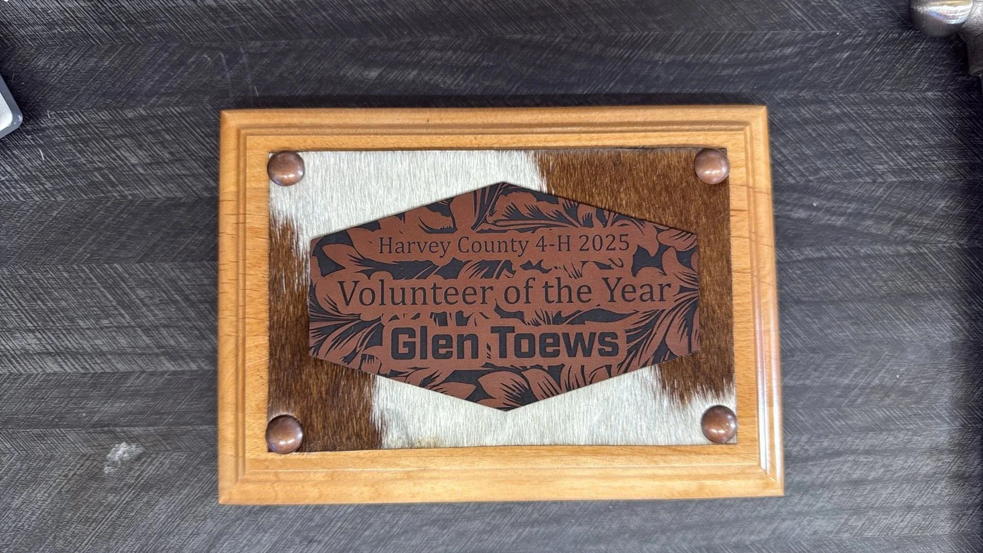 A wooden frame holding a plaque with the text "Harvey County 4-H 2025 Volunteer of the Year Glen Toews," decorated with animal fur and leather accents.