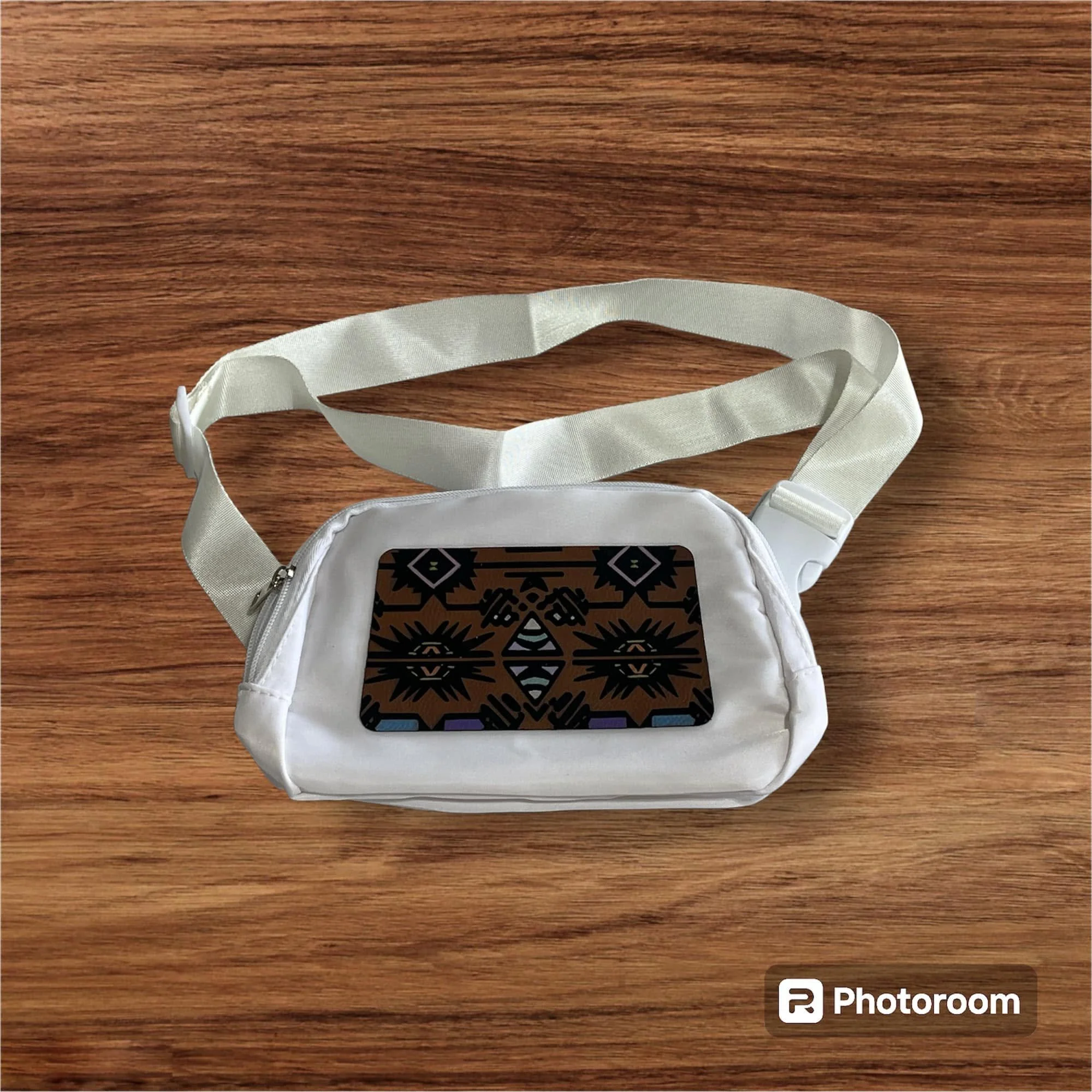 A white fabric face mask with ear loops resting on a wooden surface. The mask has a black decorative pattern on the front panel.