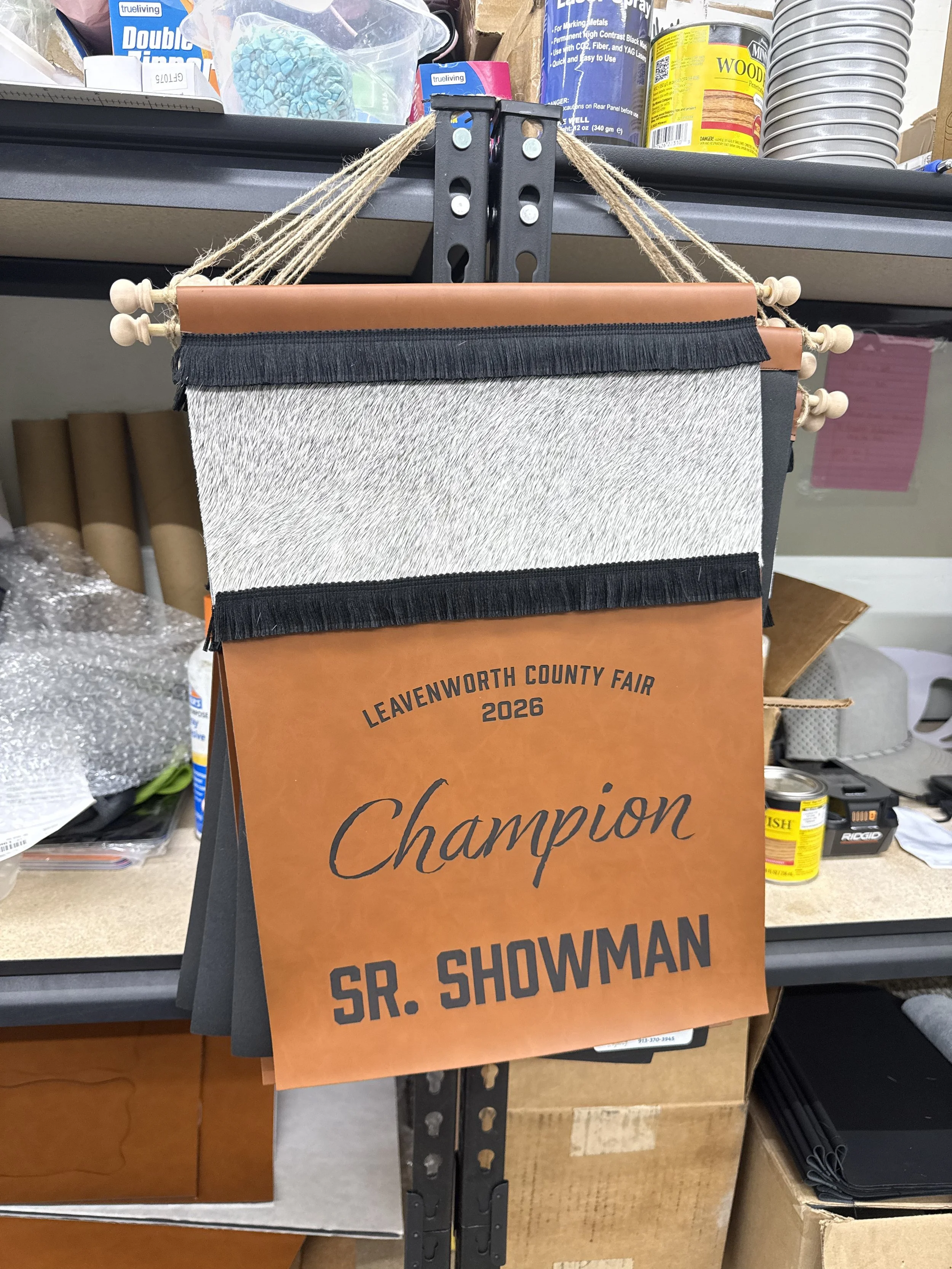 A political campaign sign for SP. Schowman, Sr. showing the message 'Leavenworth County Fair 2026 Champion' hanging on a black metal rack in a hardware store.