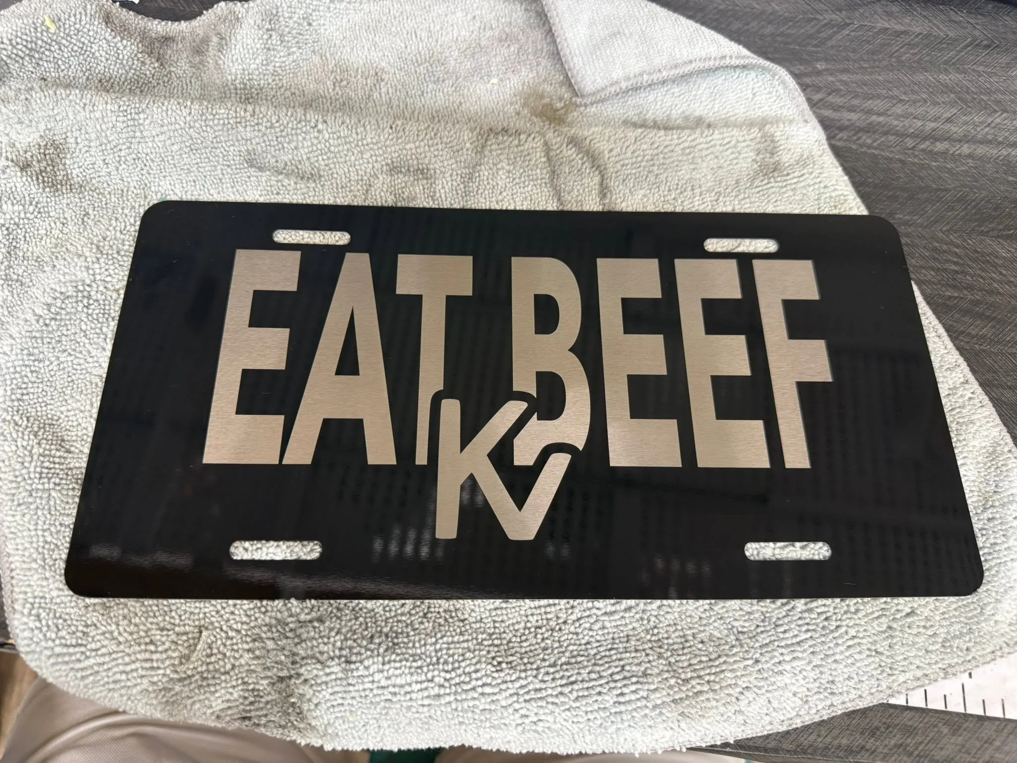 A vehicle license plate frame that says 'FAREBEE' is placed on a towel-covered surface. It has a black border and silver characters.
