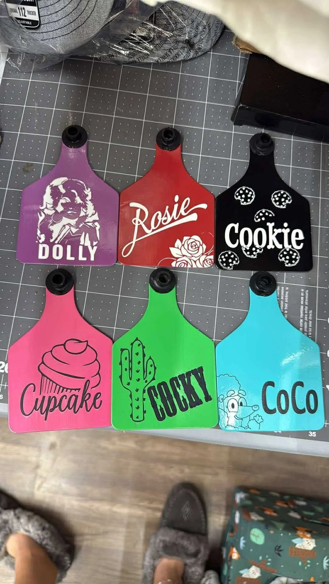 Colorful plastic bottles with black nozzles, arranged in two rows, each featuring different labels and designs. The top row includes purple labeled 'Dolly' with a girl illustration, red labeled 'Rosie' with roses, and black labeled 'Cookie' with cookie illustrations. The bottom row has pink labeled 'Cupcake' with a cupcake illustration, green labeled 'Cooky' with a cactus illustration, and blue labeled 'CoCo' with a hedgehog and tree illustration.