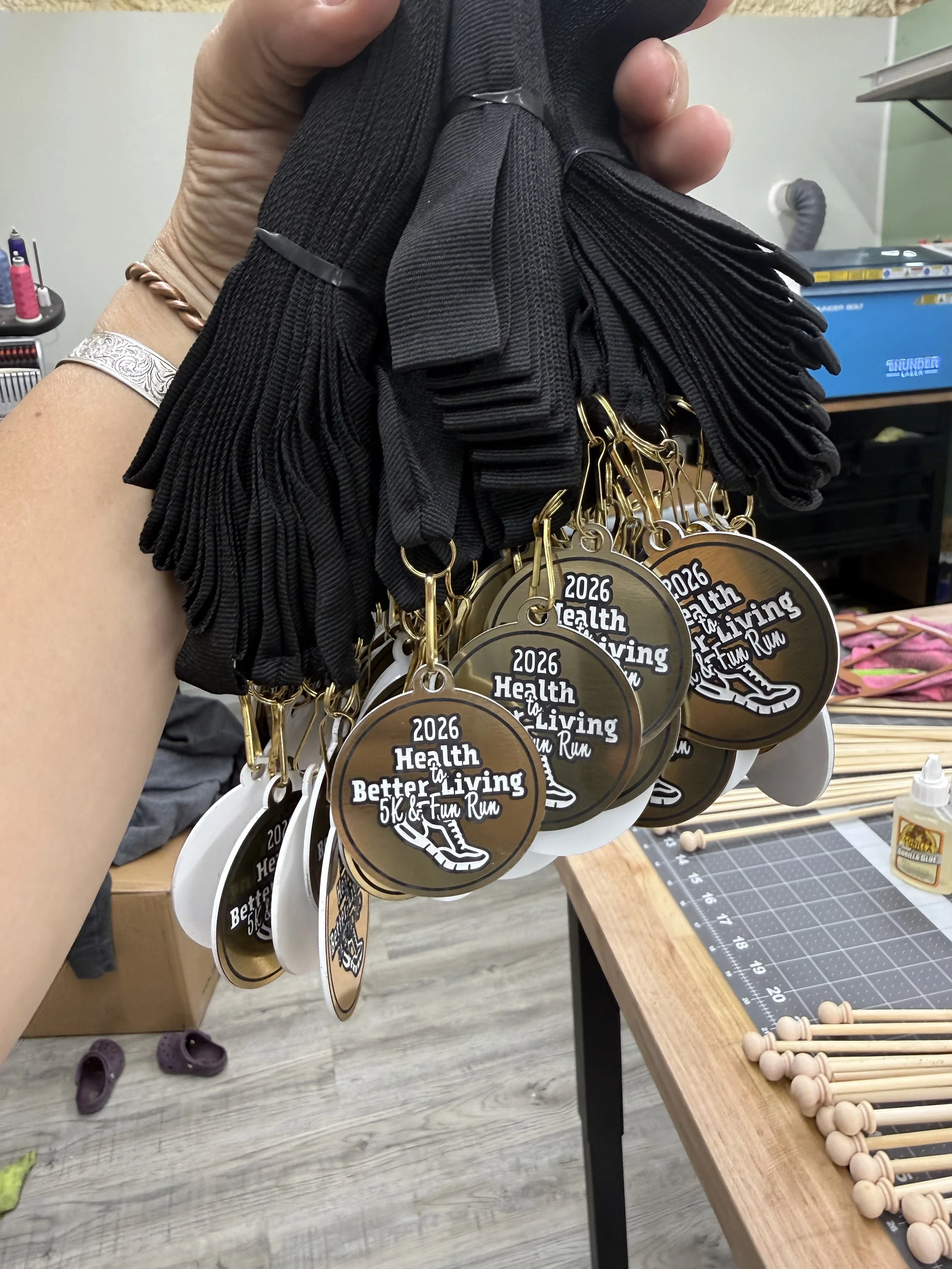 Person holding medals that read '2026 Health to Living 5K & Fun Run' with a graphic of a running shoe, medals dangling from gold clips. Nearby are wooden percussion mallets on a table.