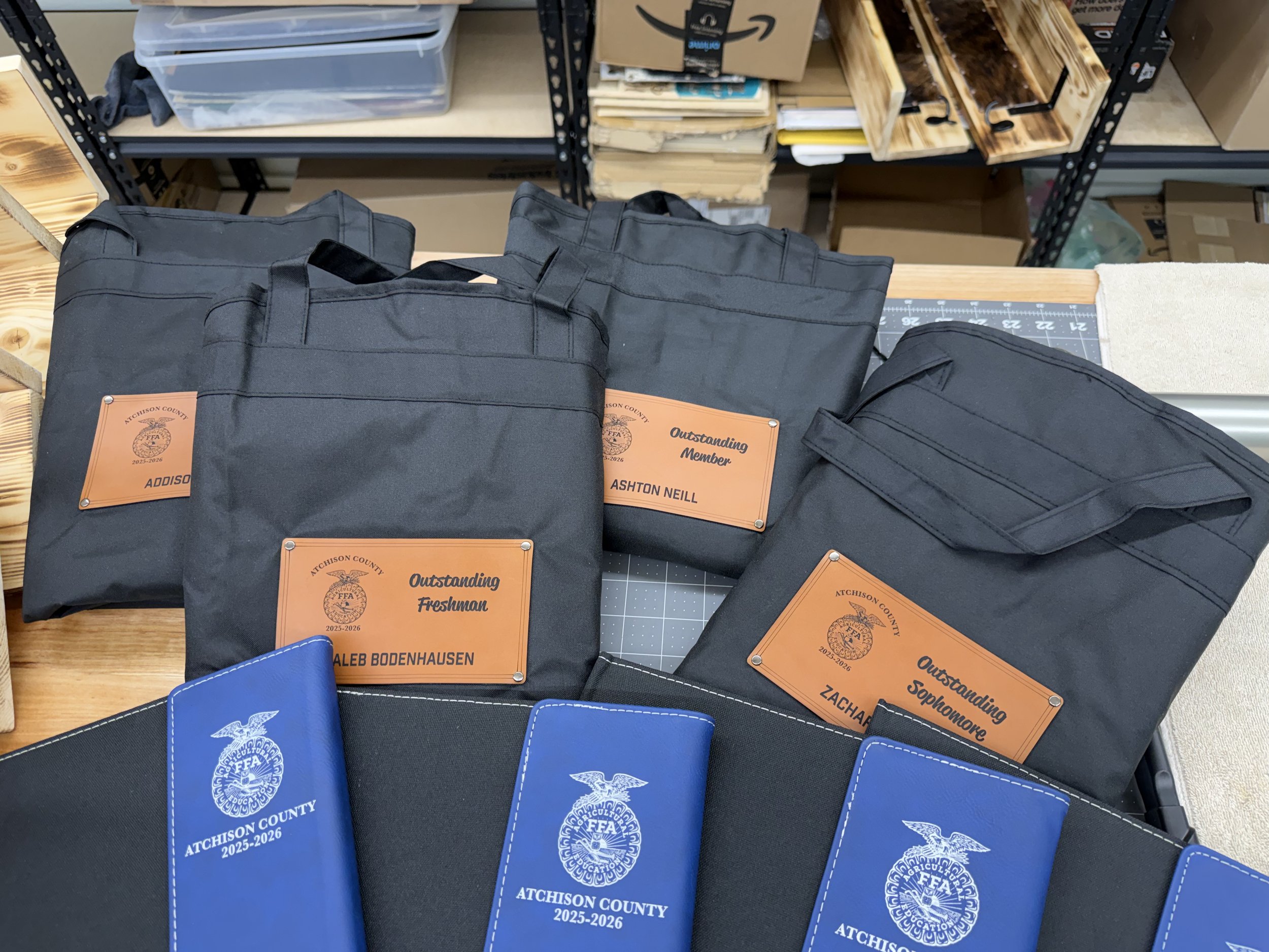 Black and blue folders with orange and blue labels that read 'Attchison County 2025-2026' and 'Outstanding Freshman' with students' names, on a workspace table.