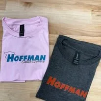 Two folded Hoffmann t-shirts, one pink and one gray, on a wooden surface.