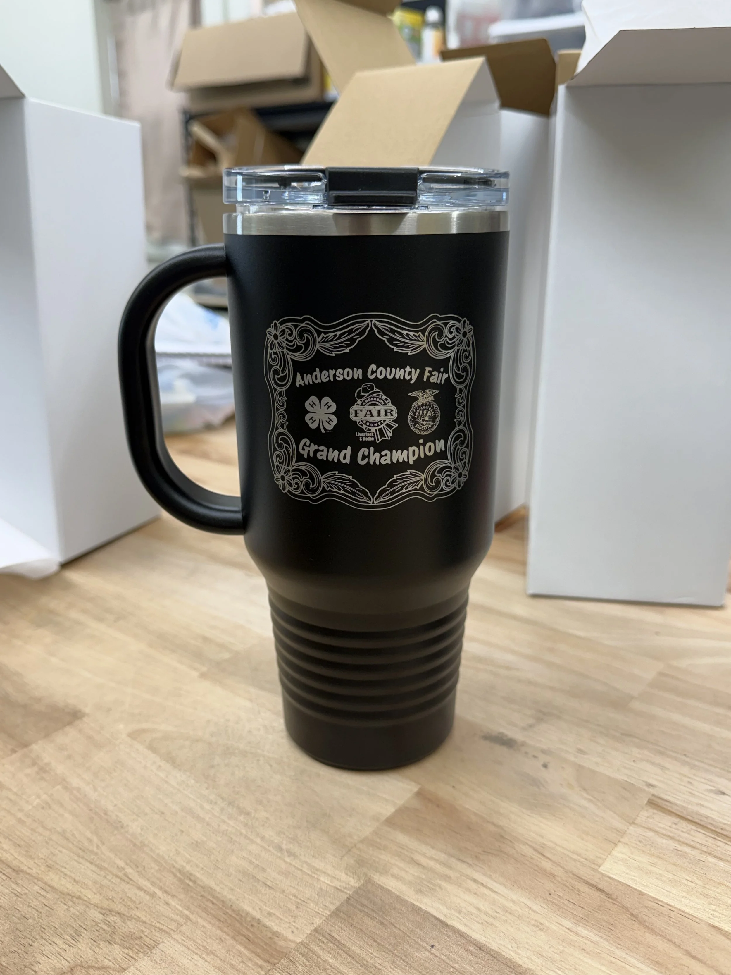 Black travel mug with a silver top and a text logo that reads 'Anderson County Fair Grand Champion' on a wooden table.
