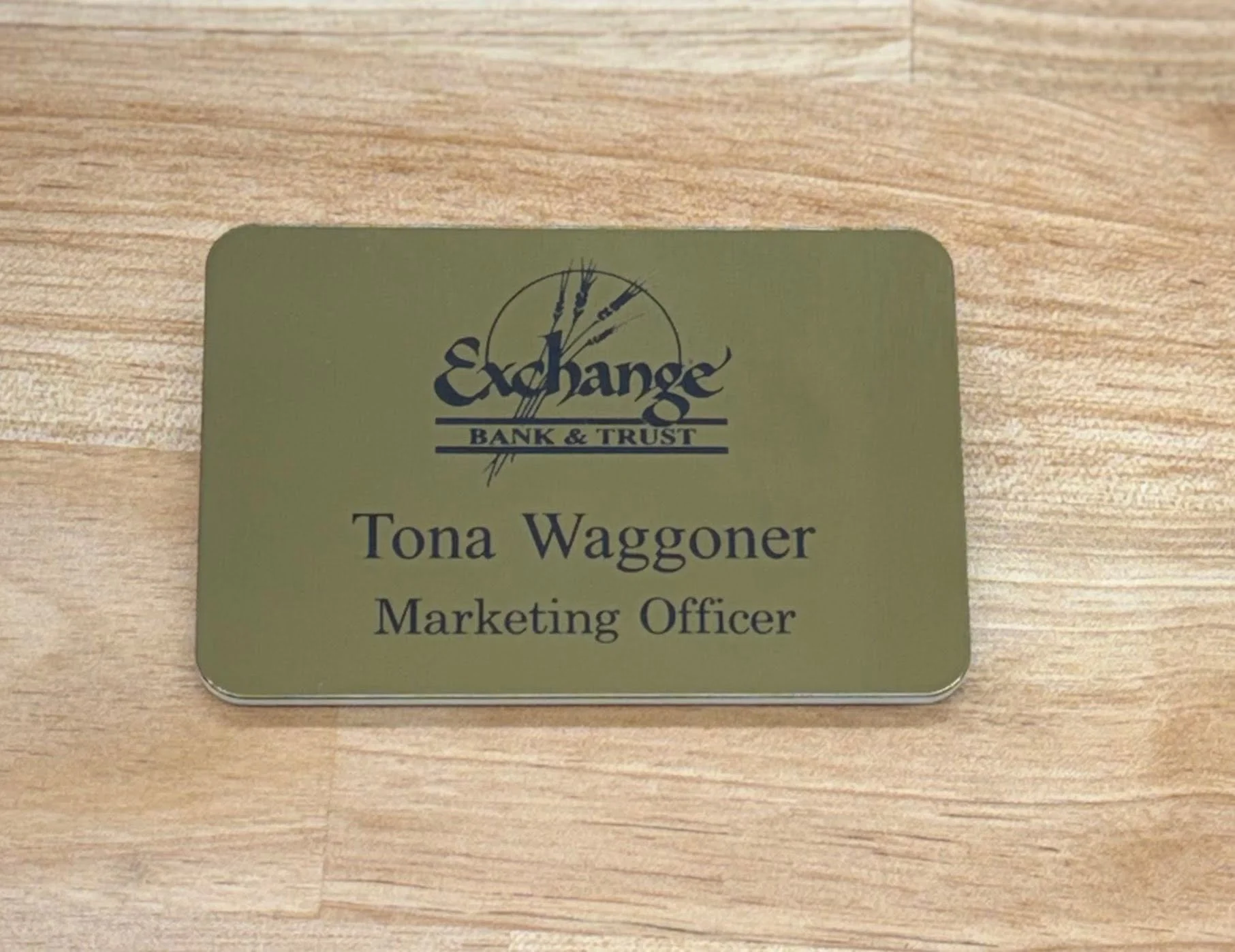 Close-up of a gold-colored badge with the logo of Exchange Bank & Trust and the name Tona Waggoner, Marketing Officer, printed below.