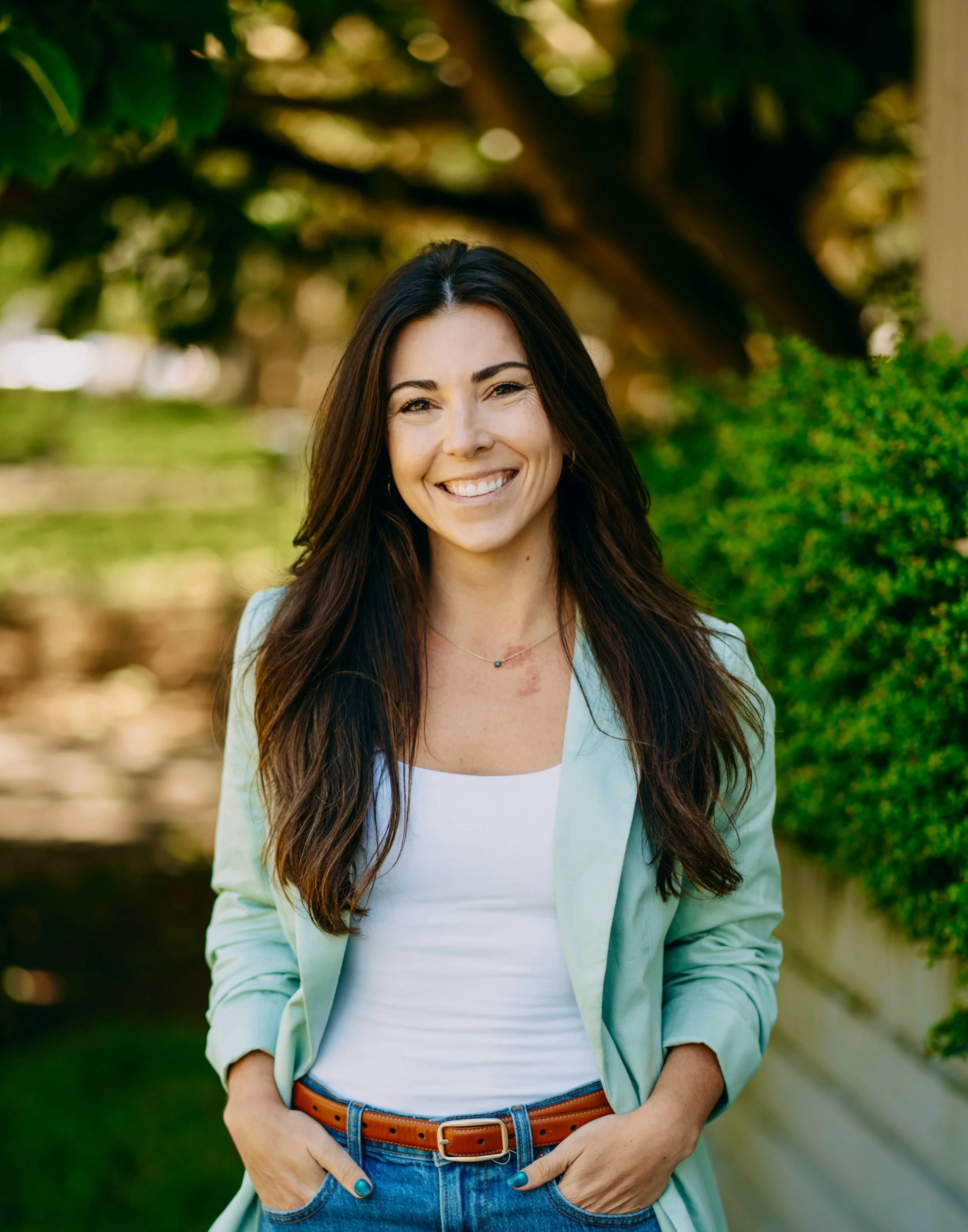 A smiling woman with long brown hair, wearing a light green blazer, white top, blue jeans, and a brown belt, standing outdoors with green bushes and trees in the background.