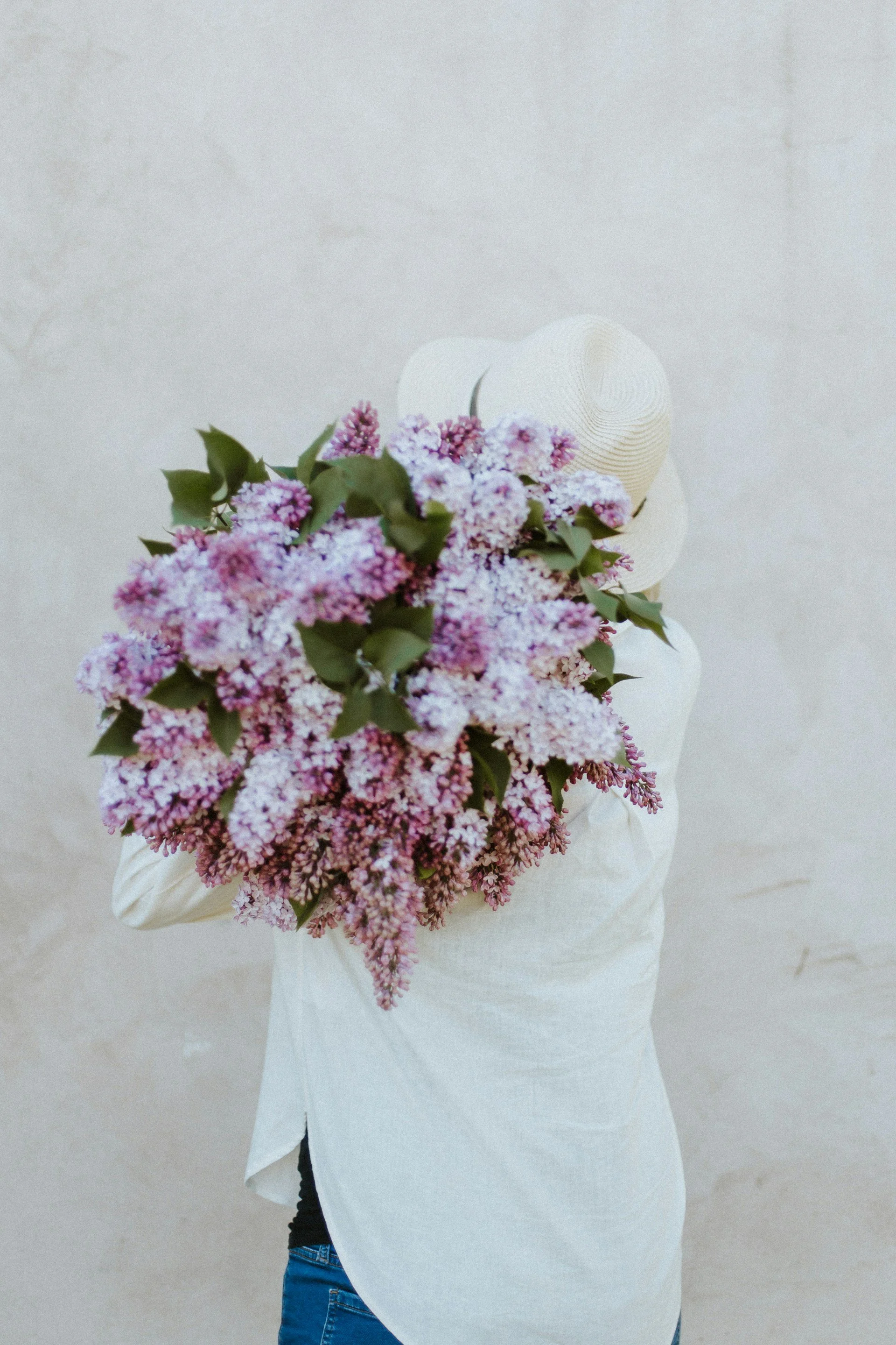 Person holding a large bouquet of pink and purple flowers, wearing a white long sleeve shirt and blue jeans, standing against a plain beige wall.