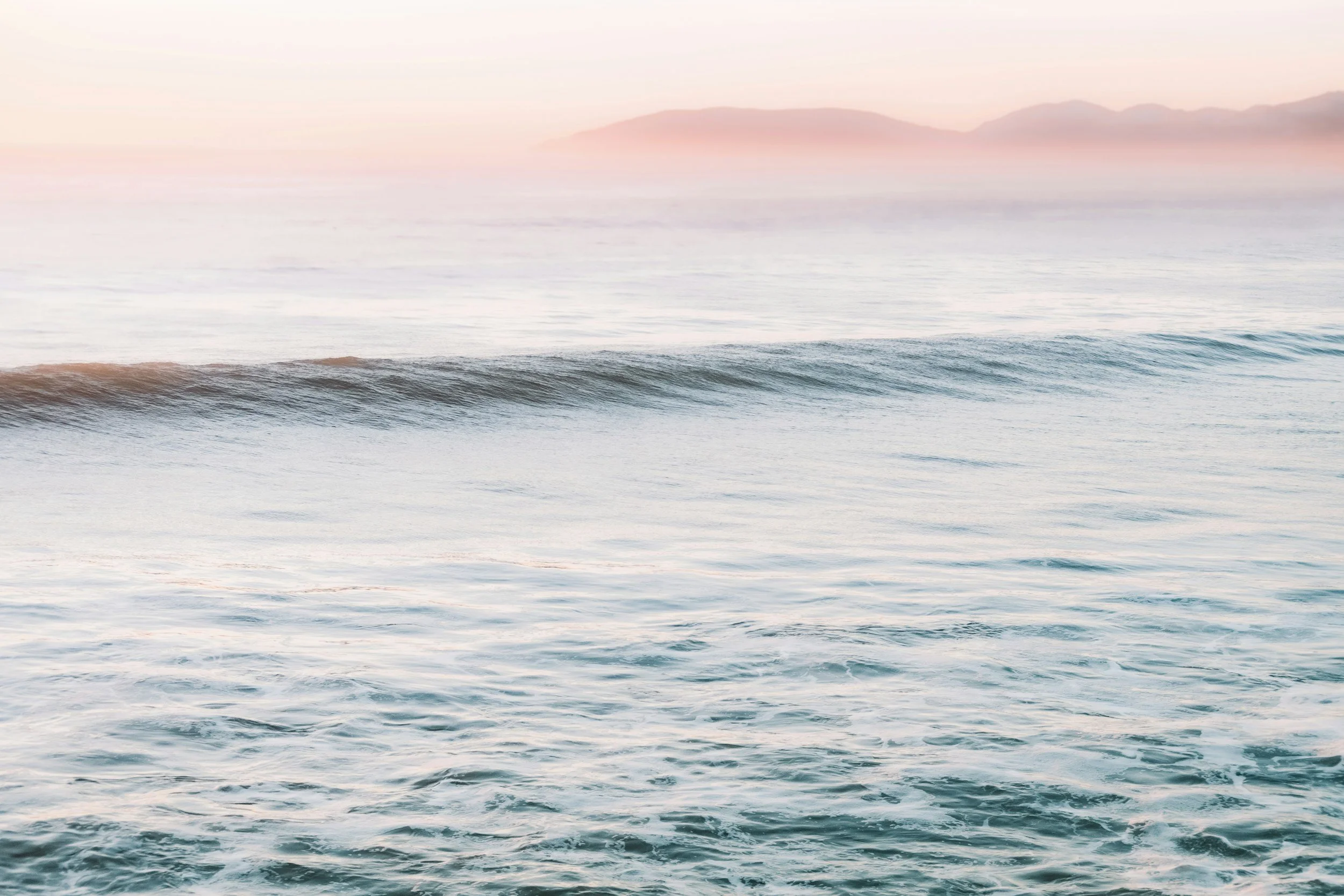 Photo of ocean waves with distant mountains and a pastel-colored sky.