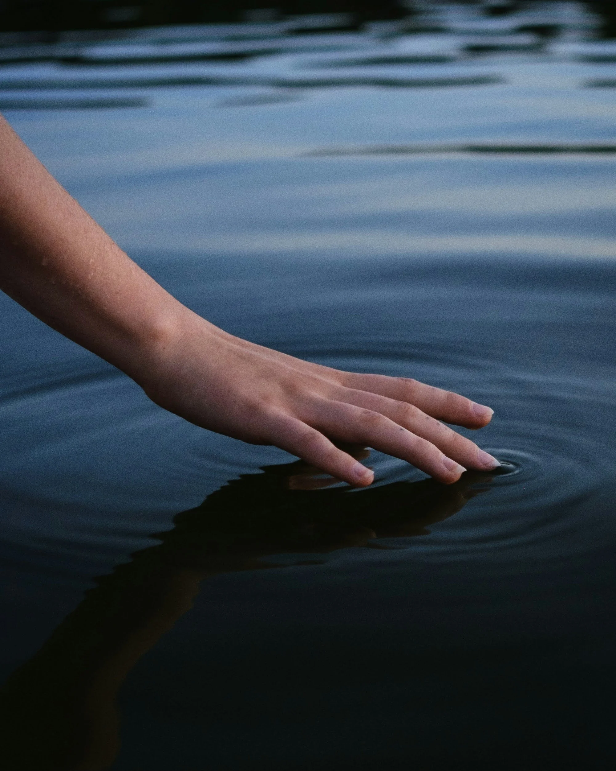 A close-up of a person's hand and fingers touching the surface of dark water, with ripples visible on the water.