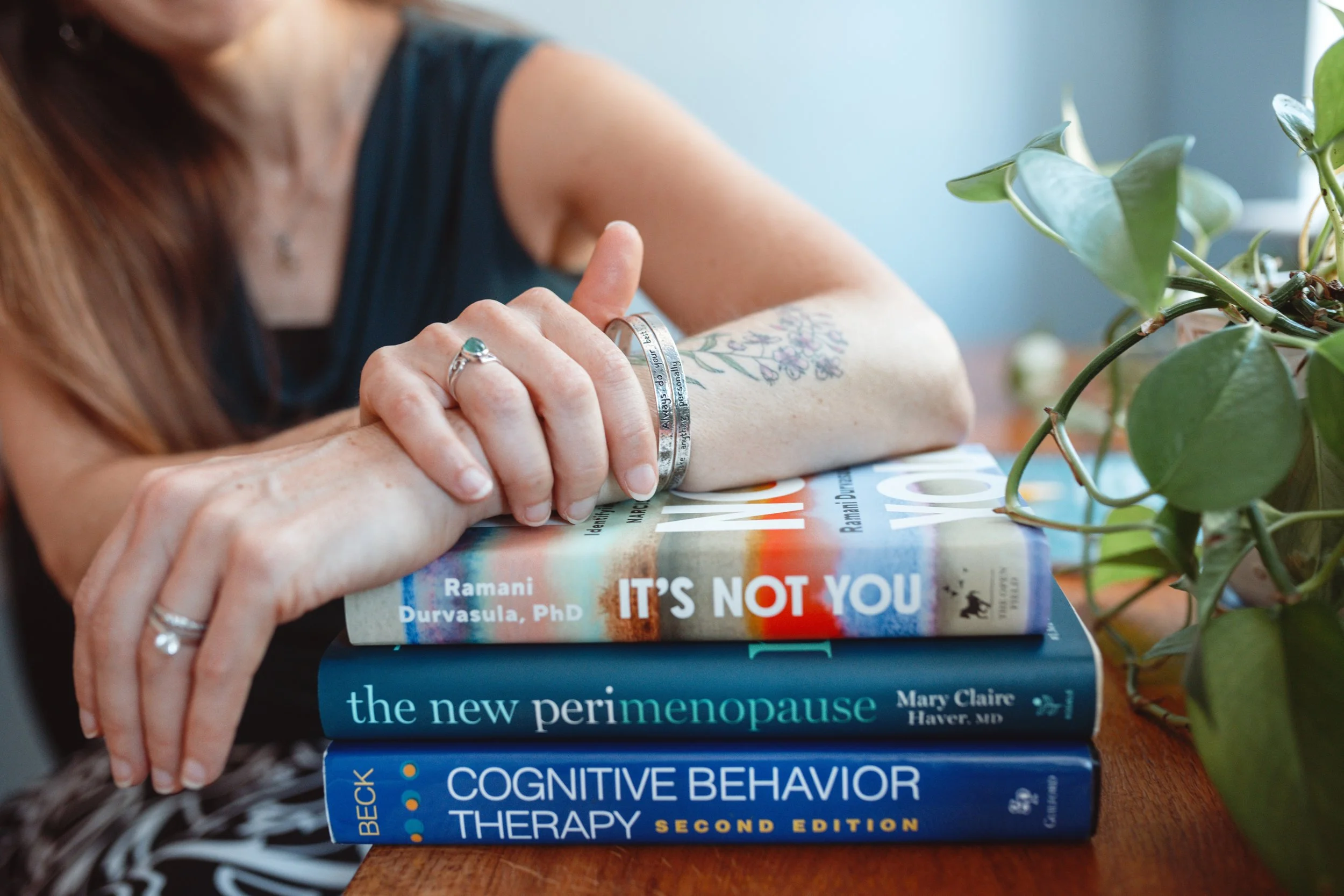 A person with long hair, wearing rings, resting their arm on a stack of books about psychology and mental health. The books include titles on menopause, cognitive behavior therapy, and mental health.