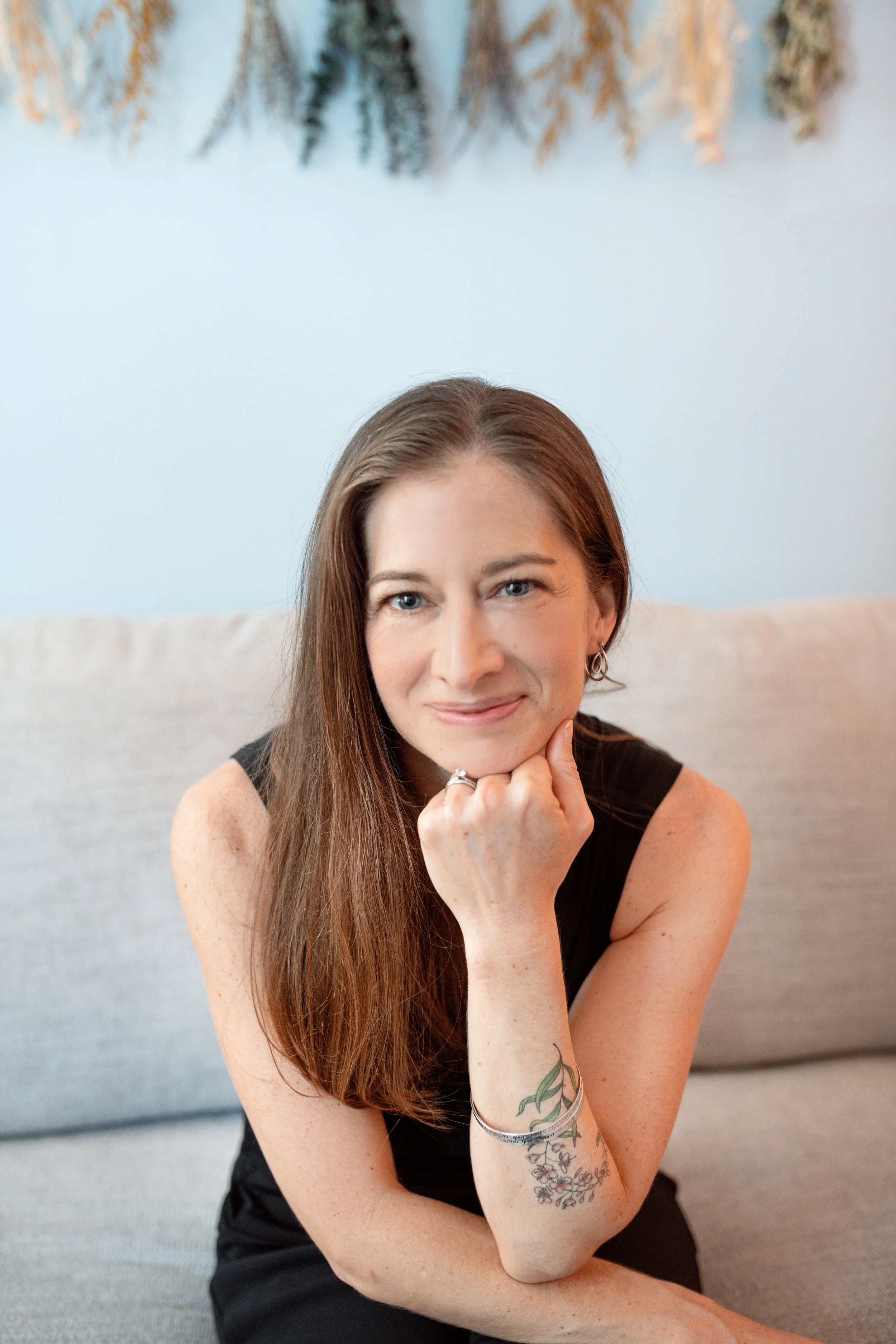 A woman with long brown hair and blue eyes, sitting on a light-colored sofa, resting her chin on her hand, wearing a black sleeveless top and earrings, with tattoos on her left forearm, in a room with a white wall and decorative dried plants or flowers hanging above her.