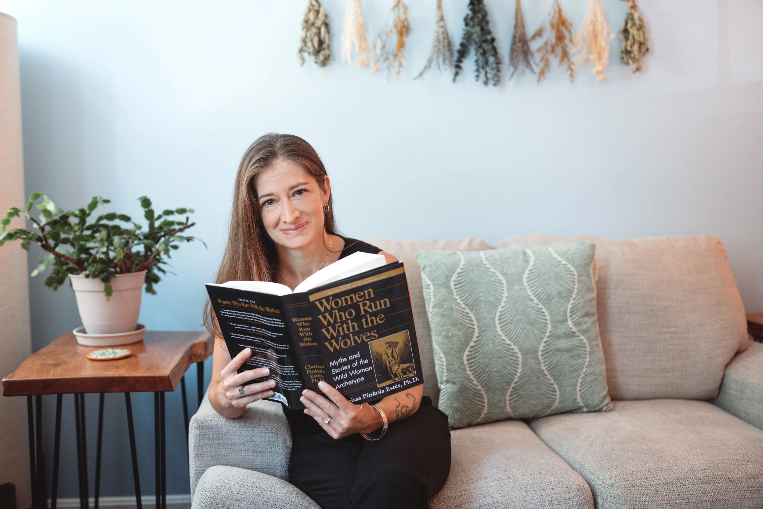 A woman with long brown hair, tattoos, and earrings sitting on a beige armchair, reading a book titled 'Women Who Run With the Wolves' in a cozy living room with a light blue wall, a decorative dried flower wall hanging, a potted plant on a wooden side table, and patterned cushions.