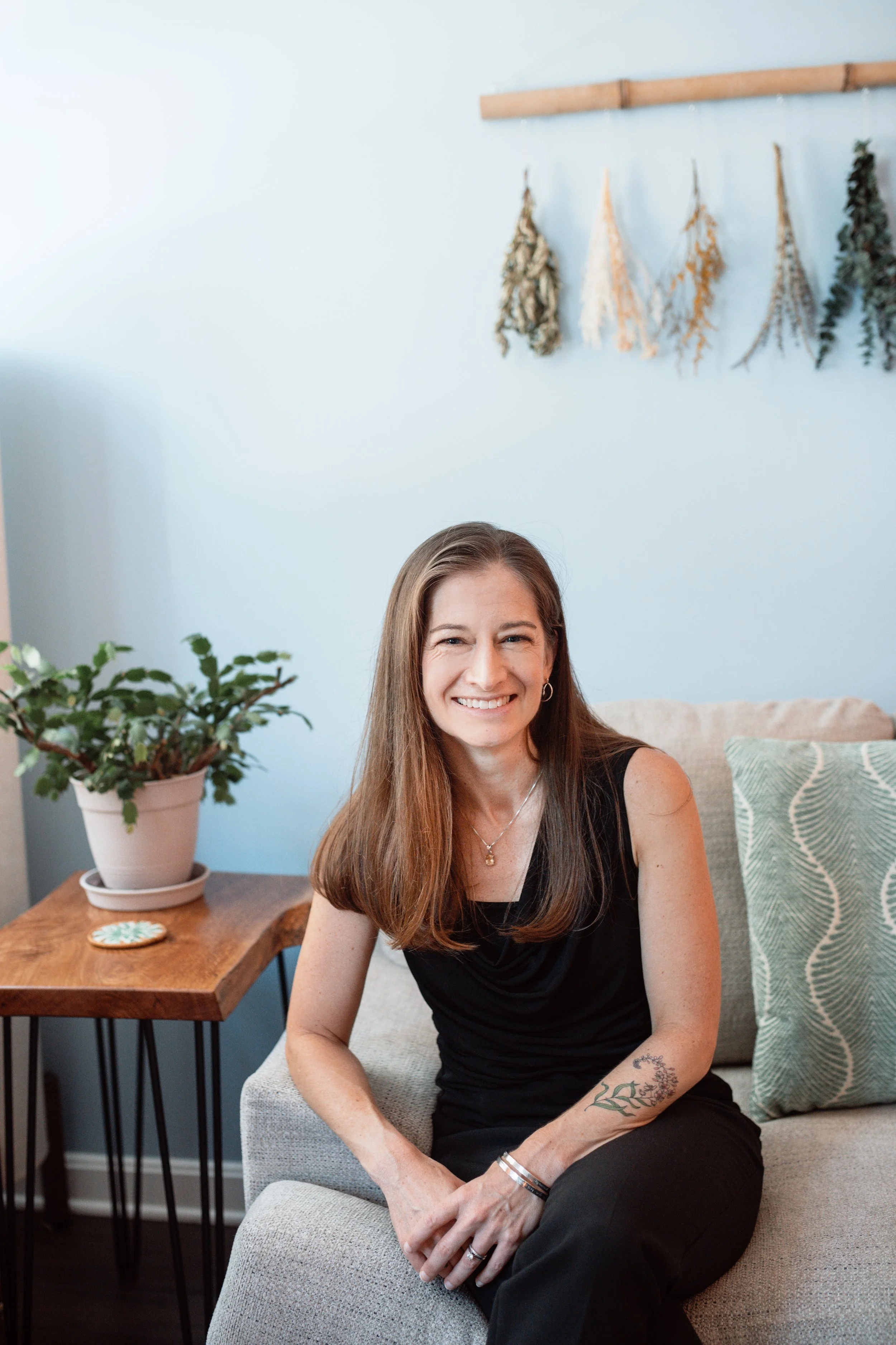 A woman with long brown hair smiling, sitting on a light-colored chair in a cozy room with blue walls, a patterned pillow, a potted plant on a wooden side table, and dried herbs hanging on the wall.