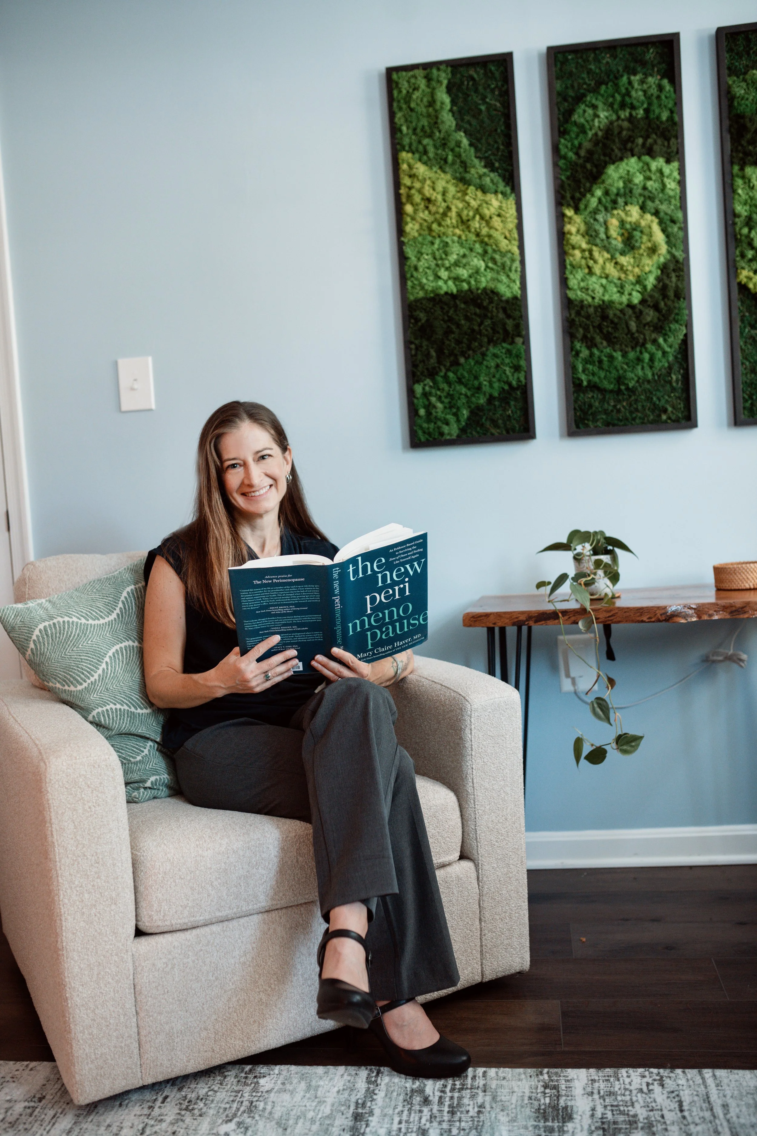 A woman with long brown hair sitting on a beige sofa, smiling and reading a book titled 'The New Permaculture Pause'. She is wearing a black top, pants, and black shoes. Behind her are three framed wall art pieces made from green moss or plants, in black frames. To her right, there is a wooden side table with green potted plants and a decorative container. The room has blue walls and dark wood flooring.