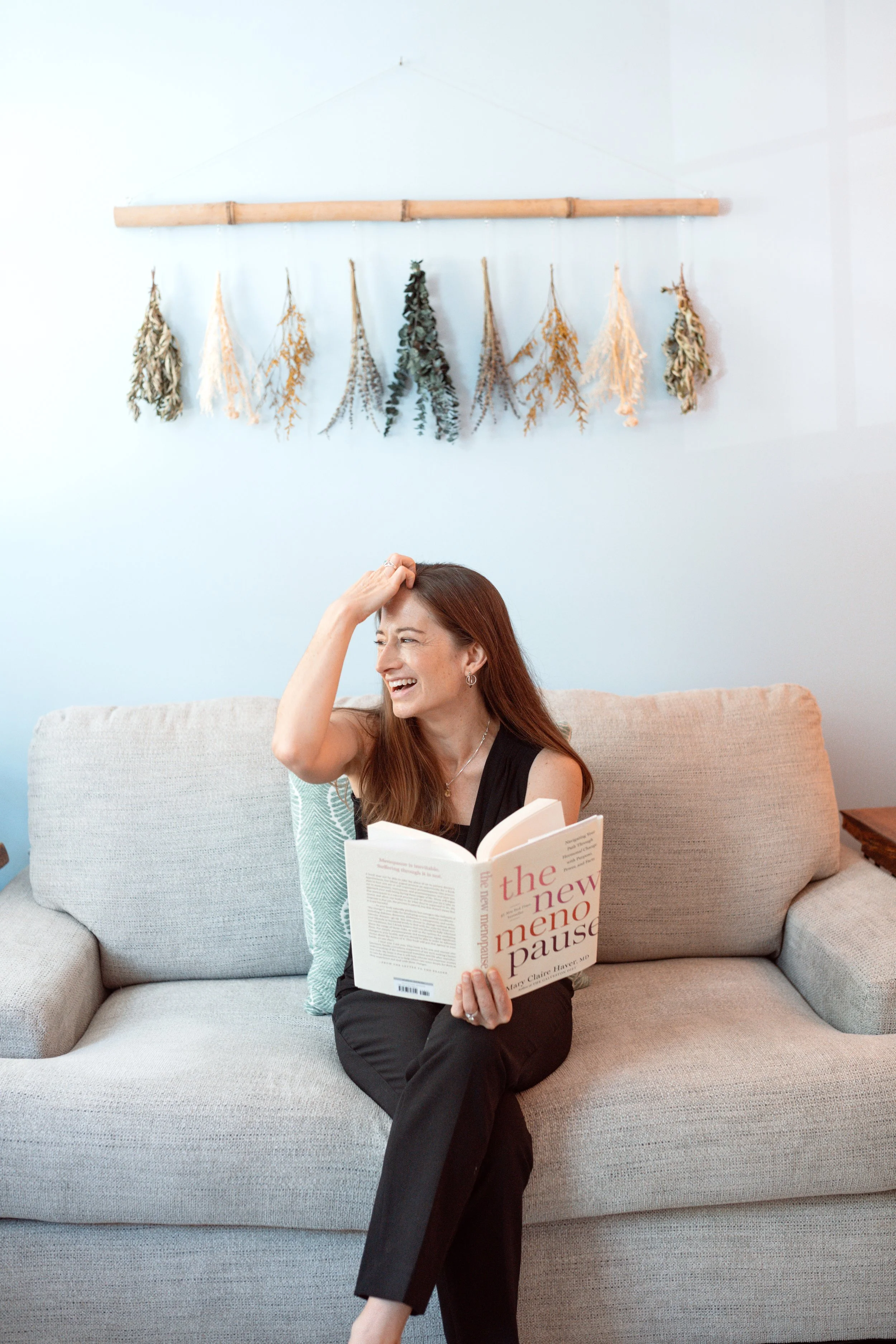 A woman with long red hair sitting on a beige couch, reading a book titled 'the new mentor pause' and smiling while touching her forehead, with a white wall and dried floral decorations hanging from a bamboo rod in the background.