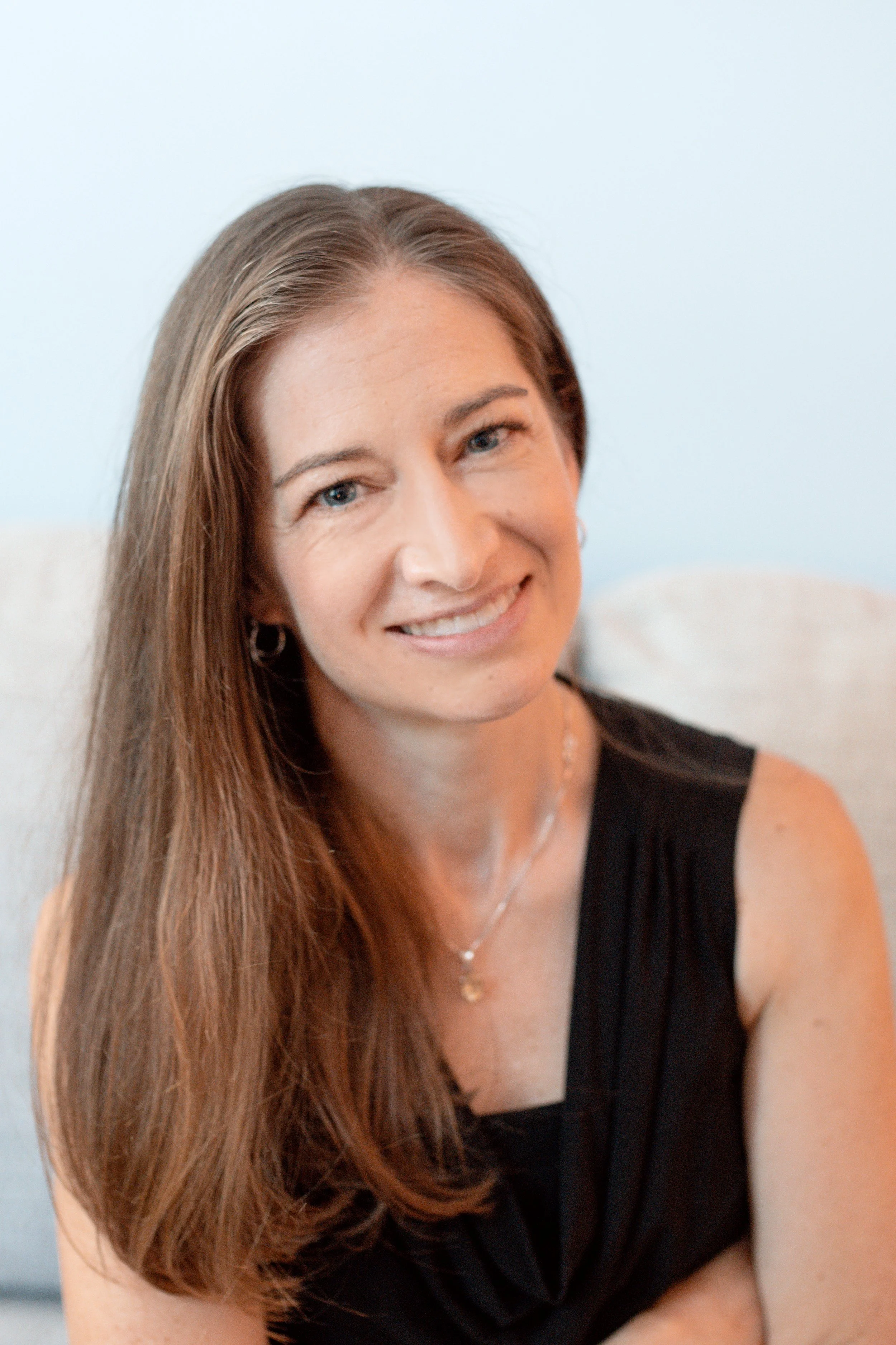 Allison Davis, LCSW-C in her therapy office in Frederick, MD, smiling and sitting indoors against a light blue wall.