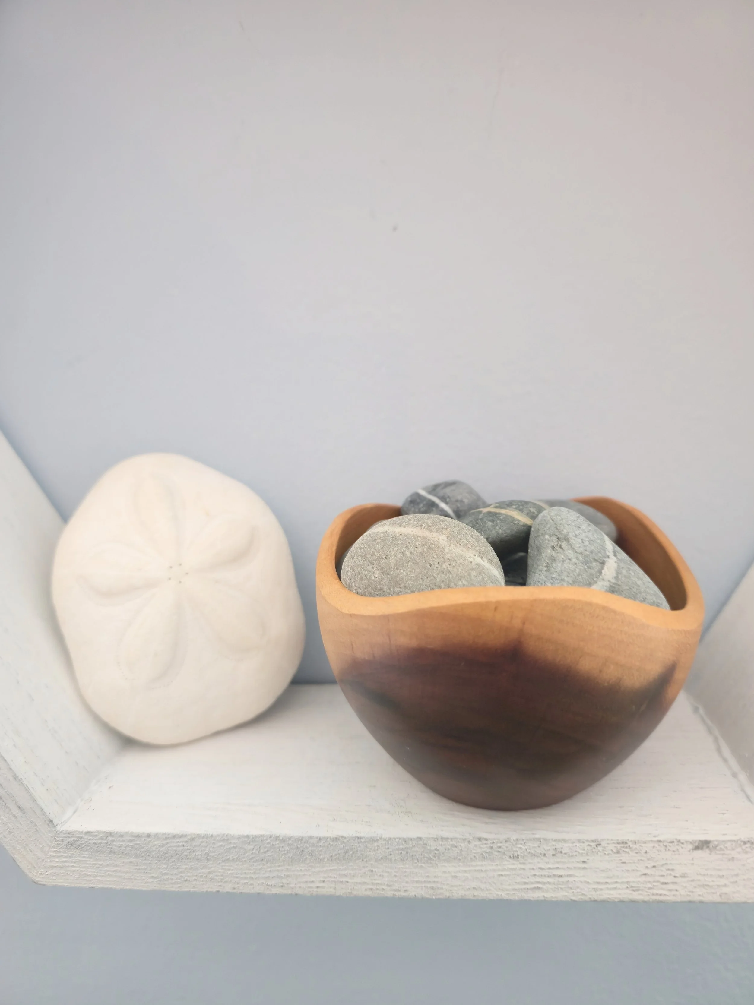 Decorative arrangement with a white seashell-shaped ornament, a small clay pot filled with smooth gray and black stones, all placed on a white wooden surface.