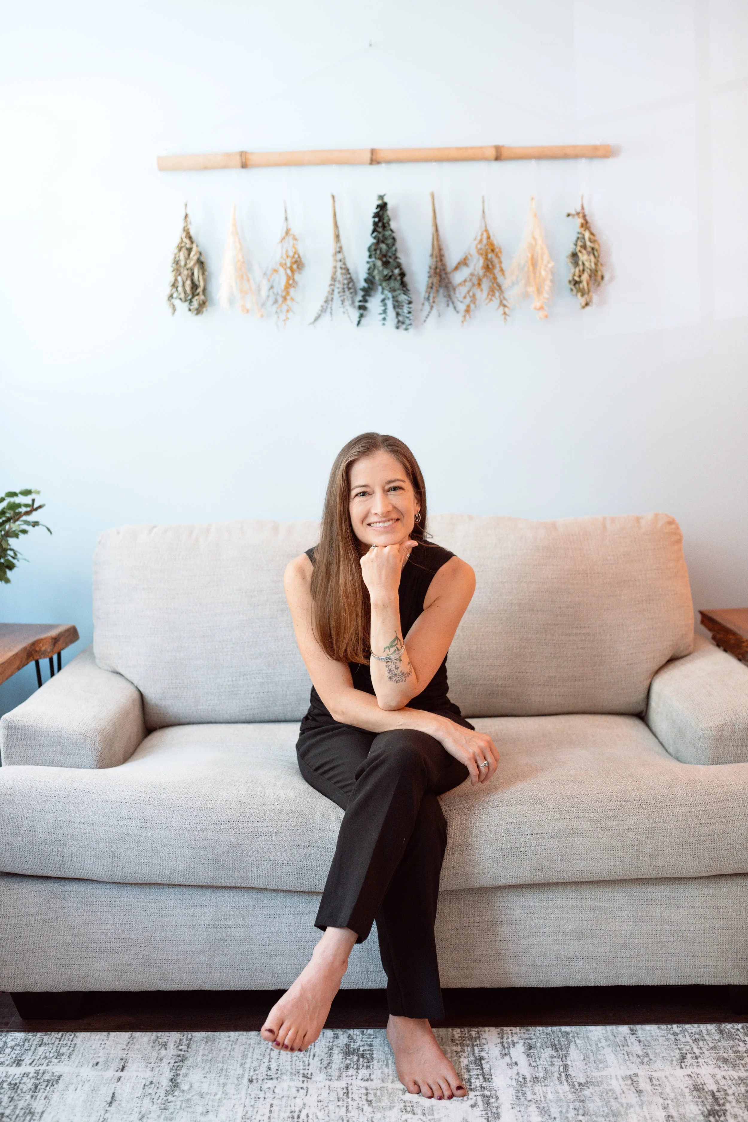 Allison Davis, LCSW-C, sitting in her therapy office in Frederick, MD on a light-colored sofa in a living room, smiling, with her hand on her chin, with a patterned rug underneath and a small potted plant on the left.