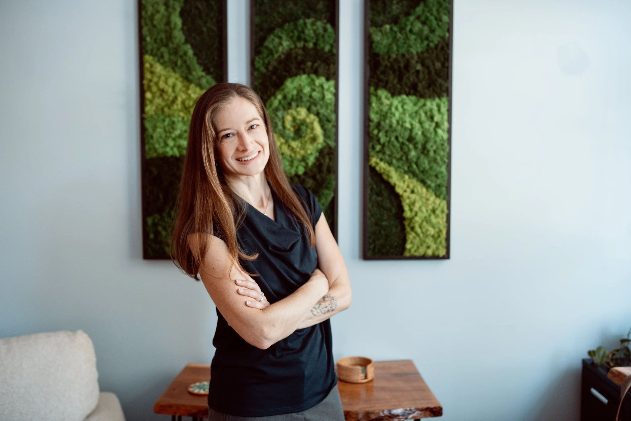 A woman with long red hair smiling and crossing her arms, standing in front of a green wall art installation inside a room with white walls and wooden furniture.