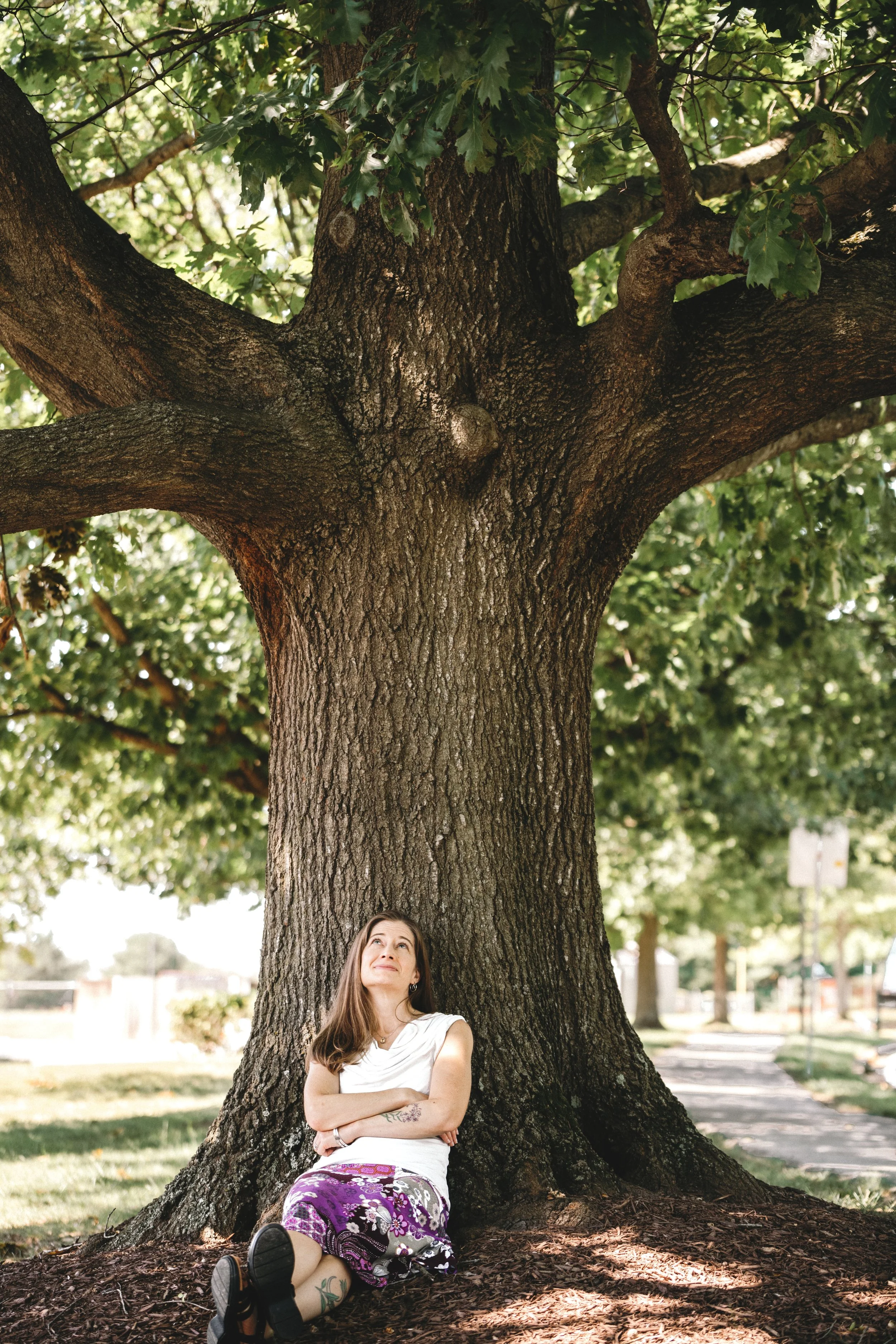 A woman with long brown hair and a white top sits with her arms crossed at the base of a large tree in a park, looking upward. The tree has a thick trunk with textured bark and broad green leaves, providing shade. There are other trees and a pathway visible in the background.
