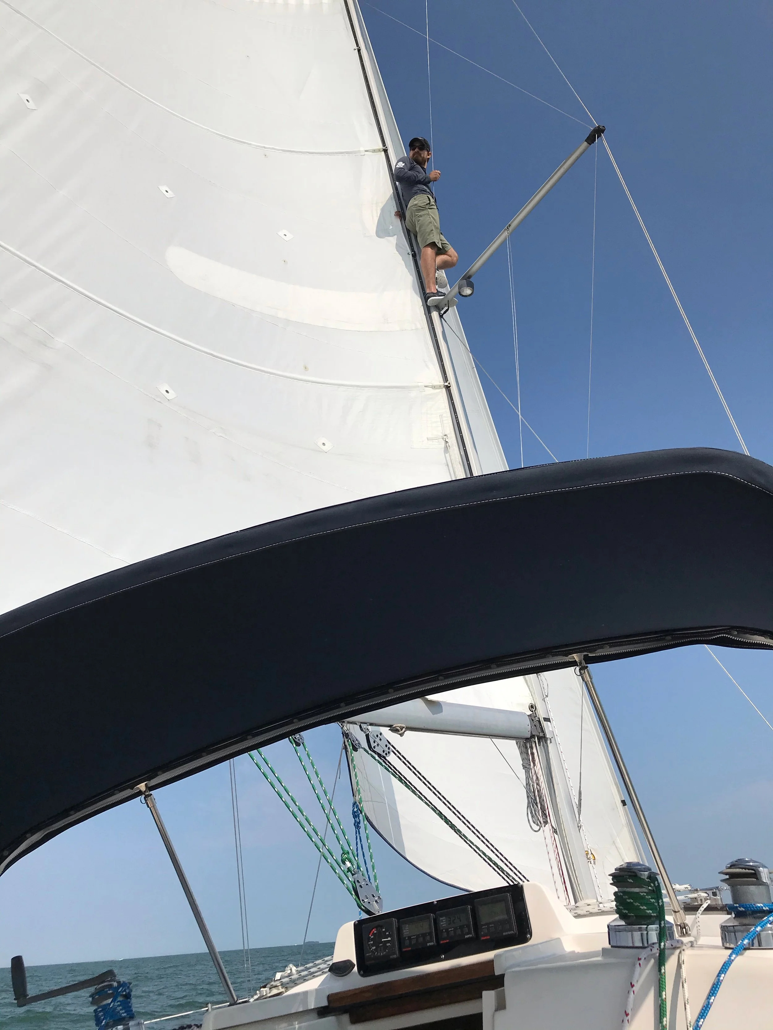 View from inside a sailboat's cockpit showing a man climbing the mast while under sail, rigging, and the sails against a clear blue sky and open water.
