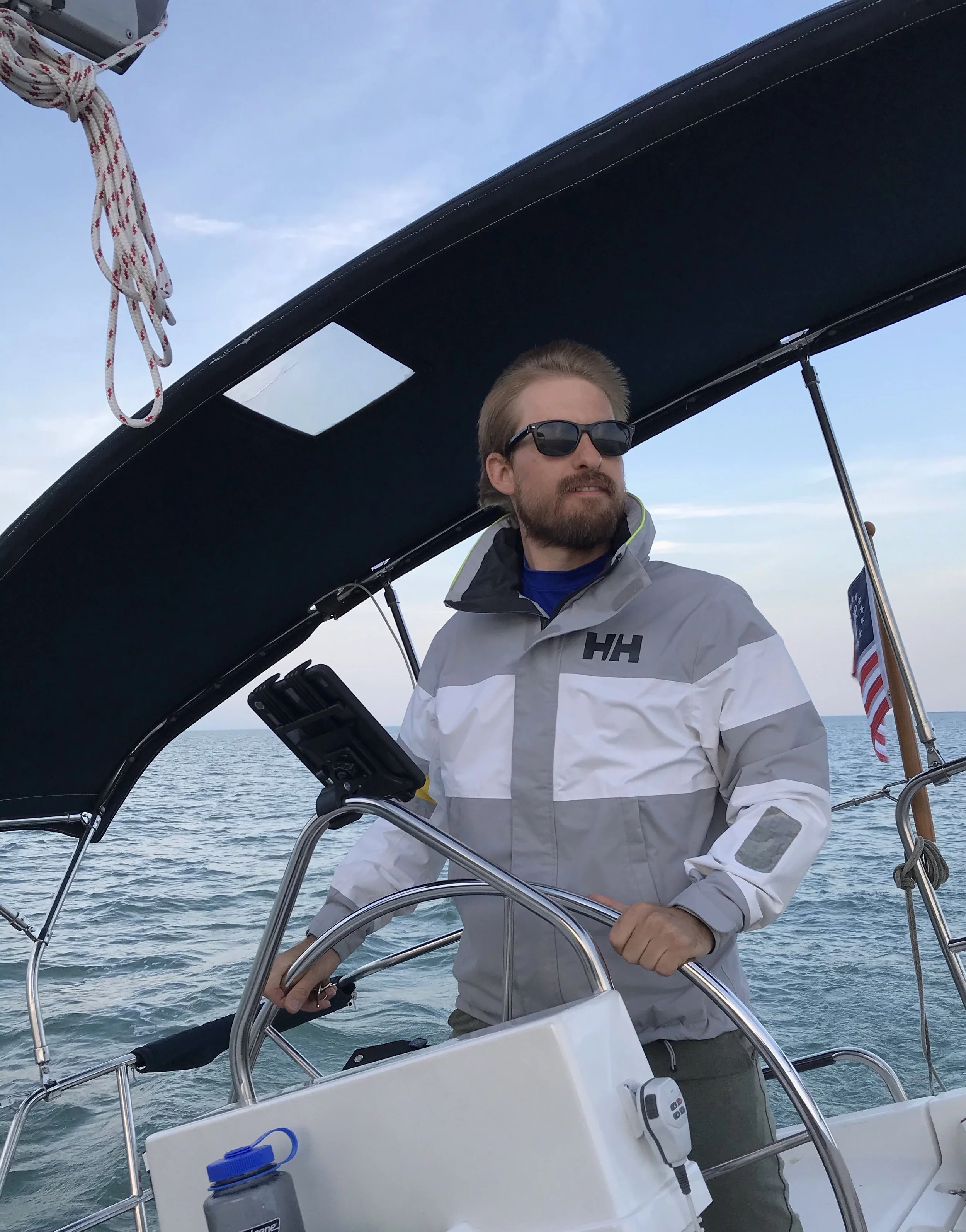 A man with a beard and sunglasses steering a sailboat on the water during the daytime, with an American flag in the background.