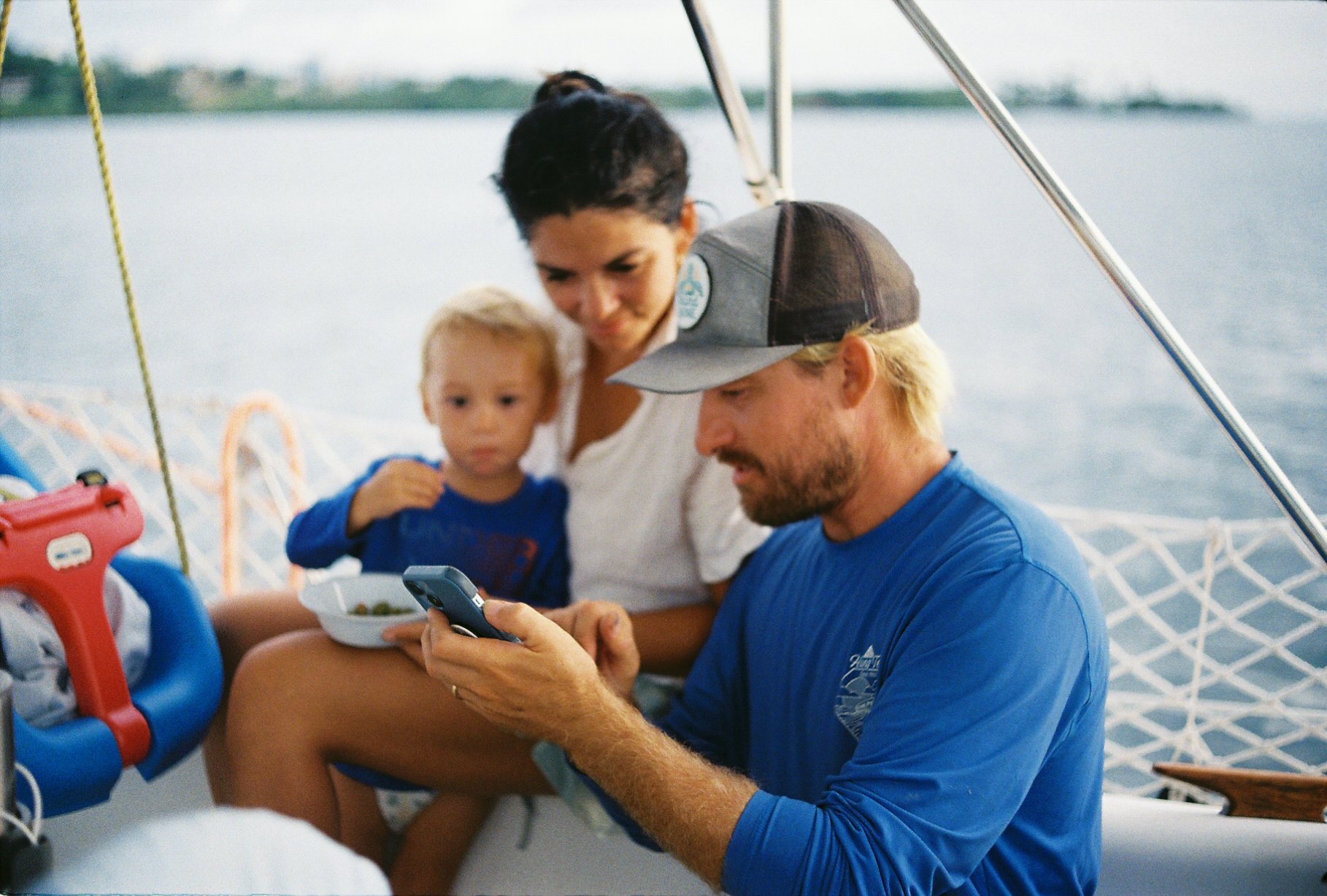 A family of three on a boat with a body of water and city skyline in the background. The man in the foreground, wearing a blue shirt and a baseball cap, is looking at a smartphone. A woman and a young child sit behind him, looking at the phone together, with the woman holding the child