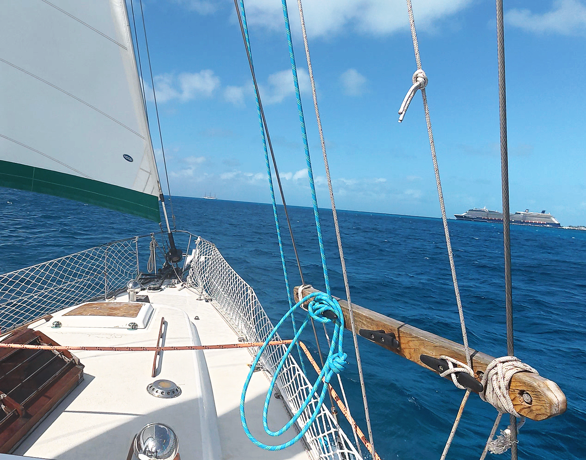View from a sailboat showing the boat's deck with various ropes, a wooden handrail, and part of a sail. In the background, the open ocean with active cruise ships under a partly cloudy sky.