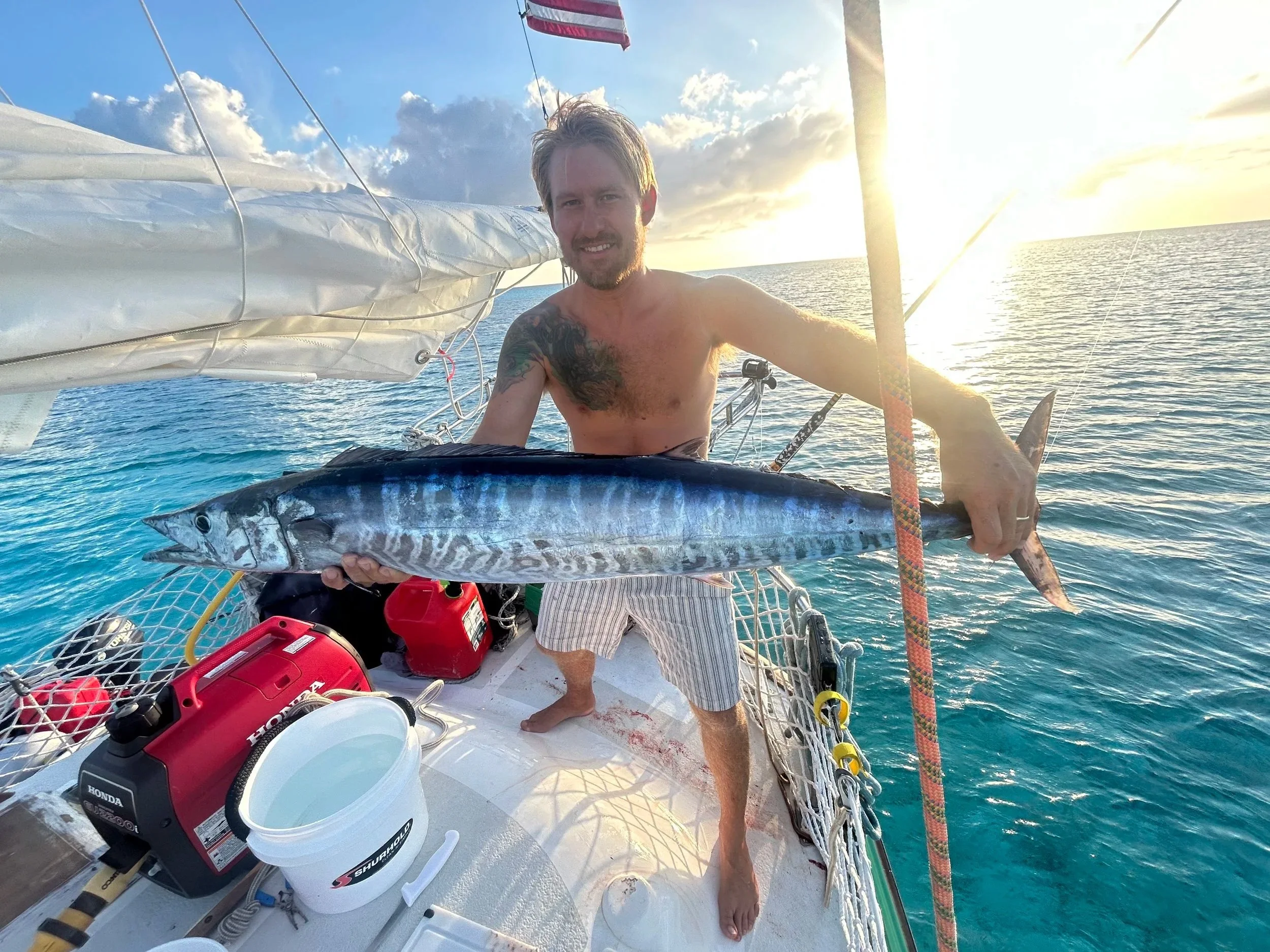 A man with a beard and tattoos, wearing striped shorts, is on a boat holding a large fish. The boat has various equipment including a red Honda generator and a white bucket. The water appears calm with the horizon and sky visible in the background, during sunset.
