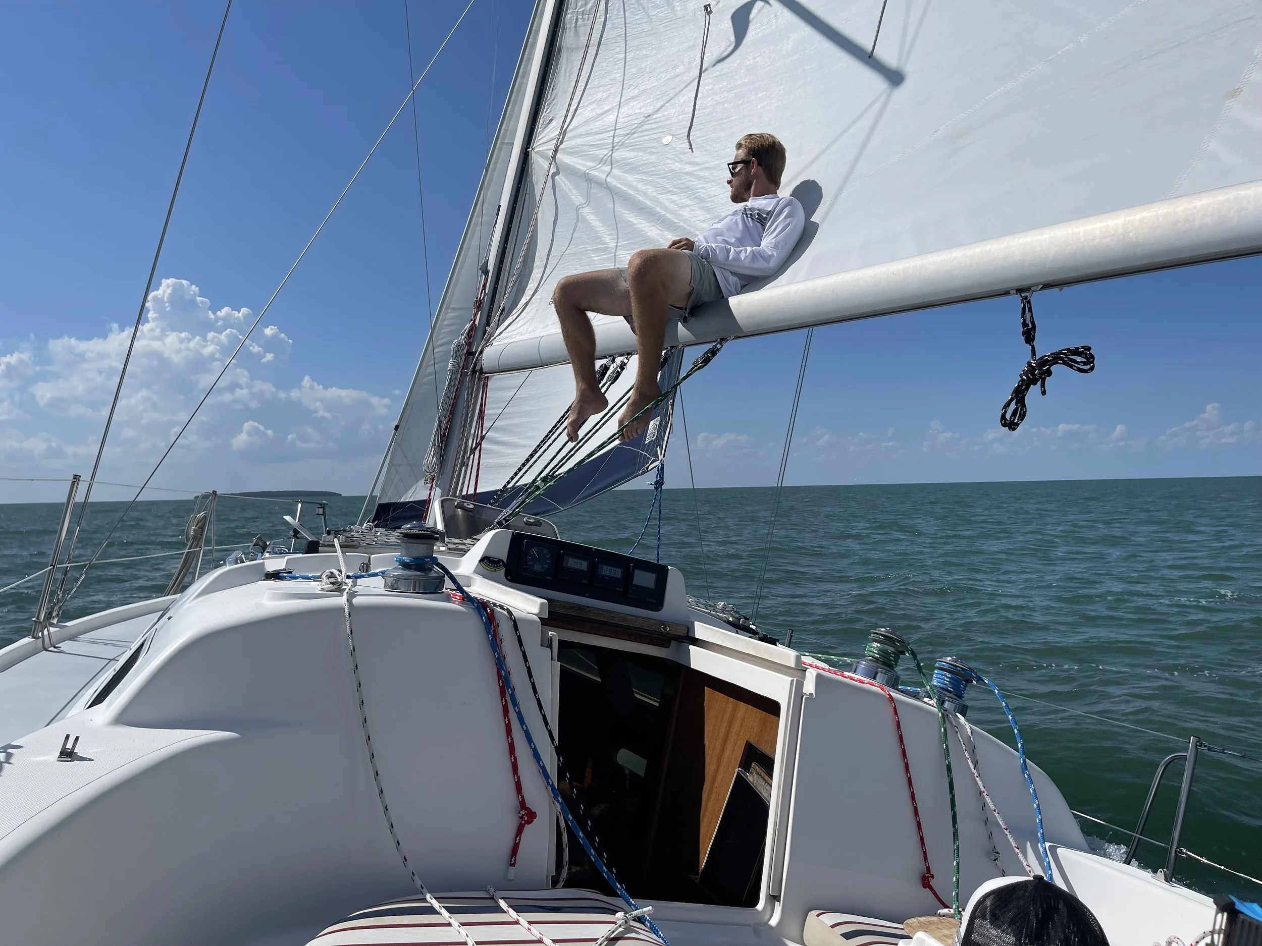 Man relaxing on the boom and sail of a sailboat under a clear blue sky with some clouds, out on the water.
