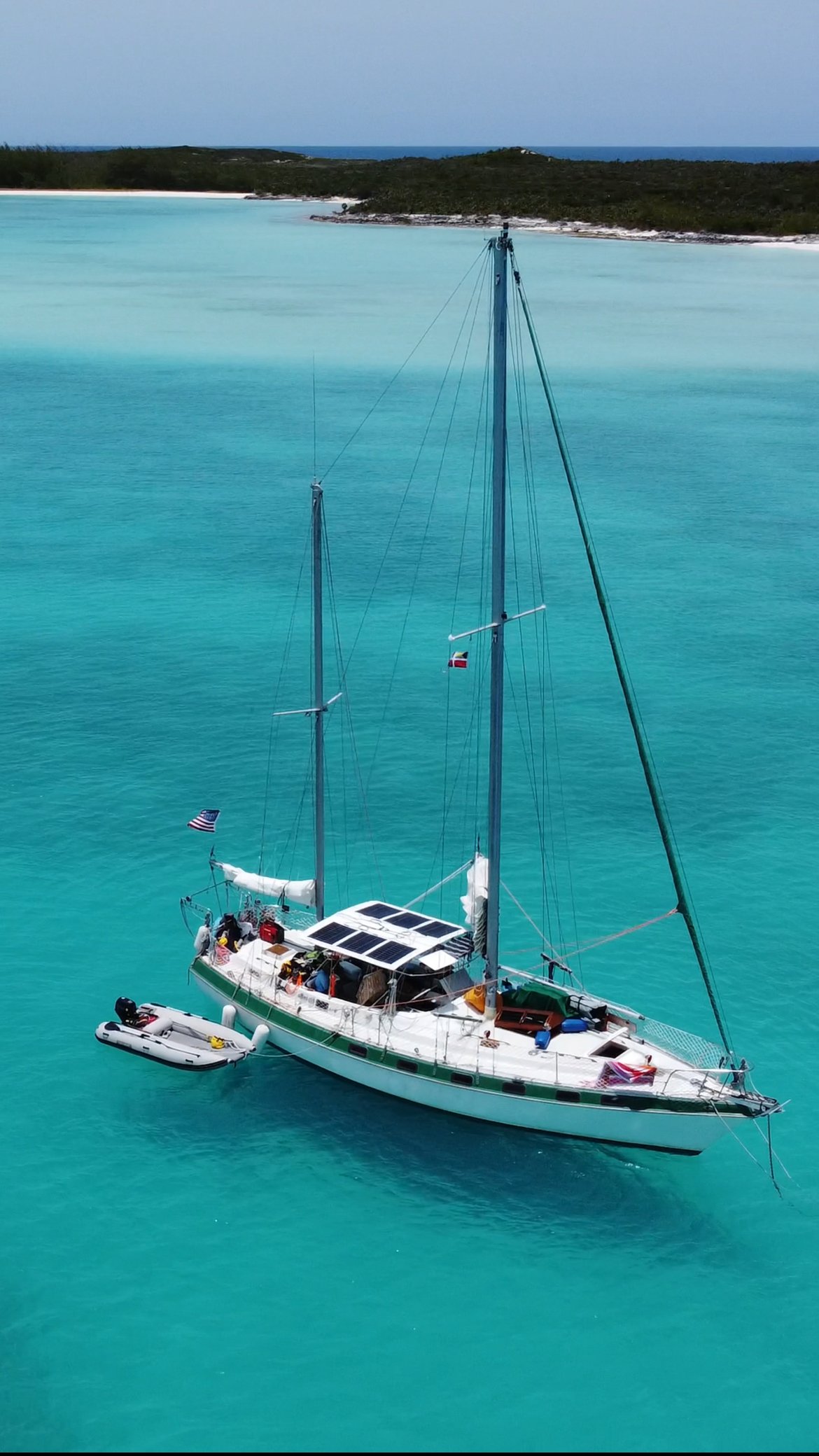 A sailboat with solar panels on the roof floating in clear turquoise water near a shoreline with greenery in the background.
