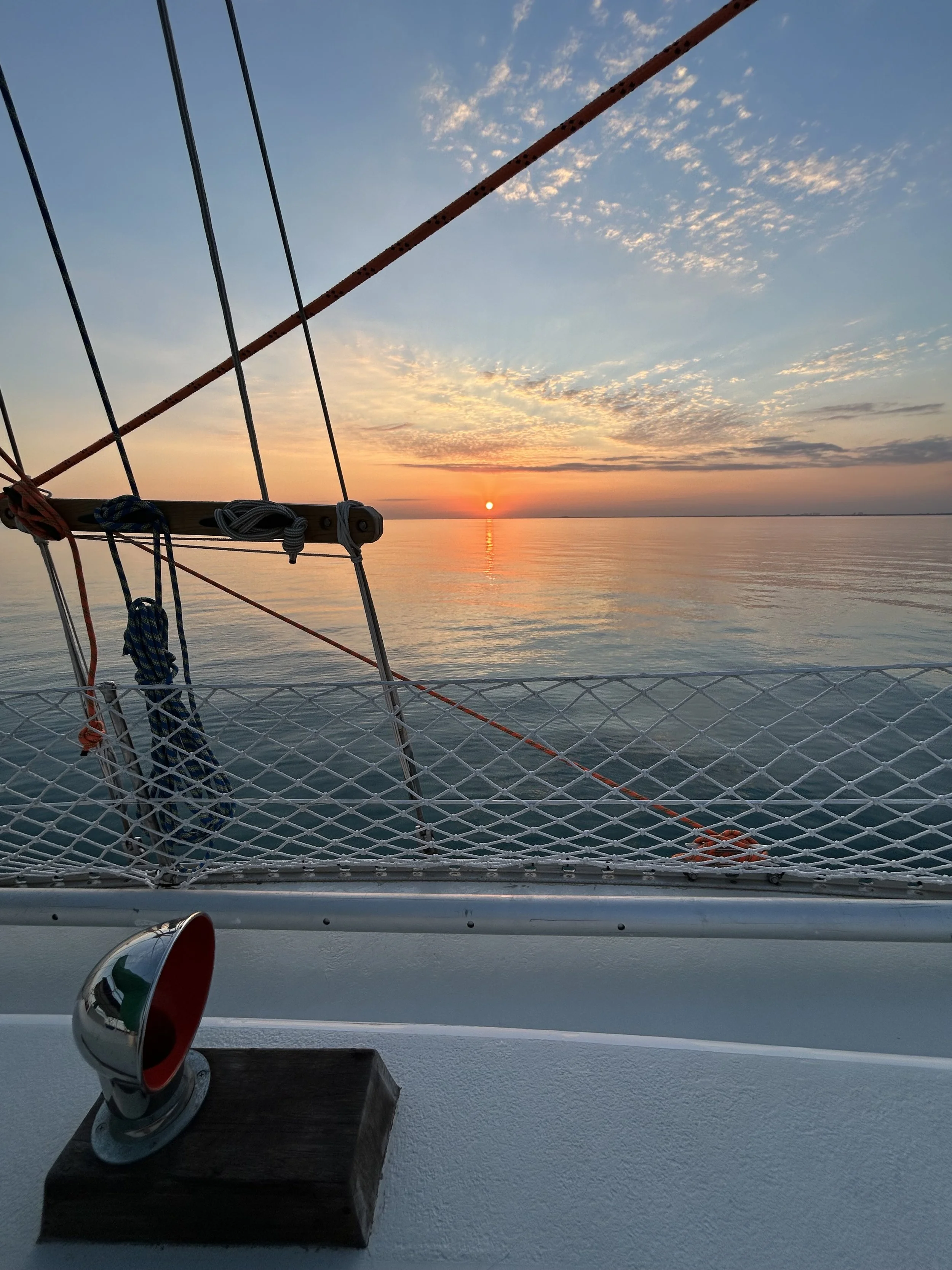 Sunset view from a boat, with a fishing rod, glasses, and a coffee mug on a table, with the sky filled with clouds and the water reflecting the sunset.