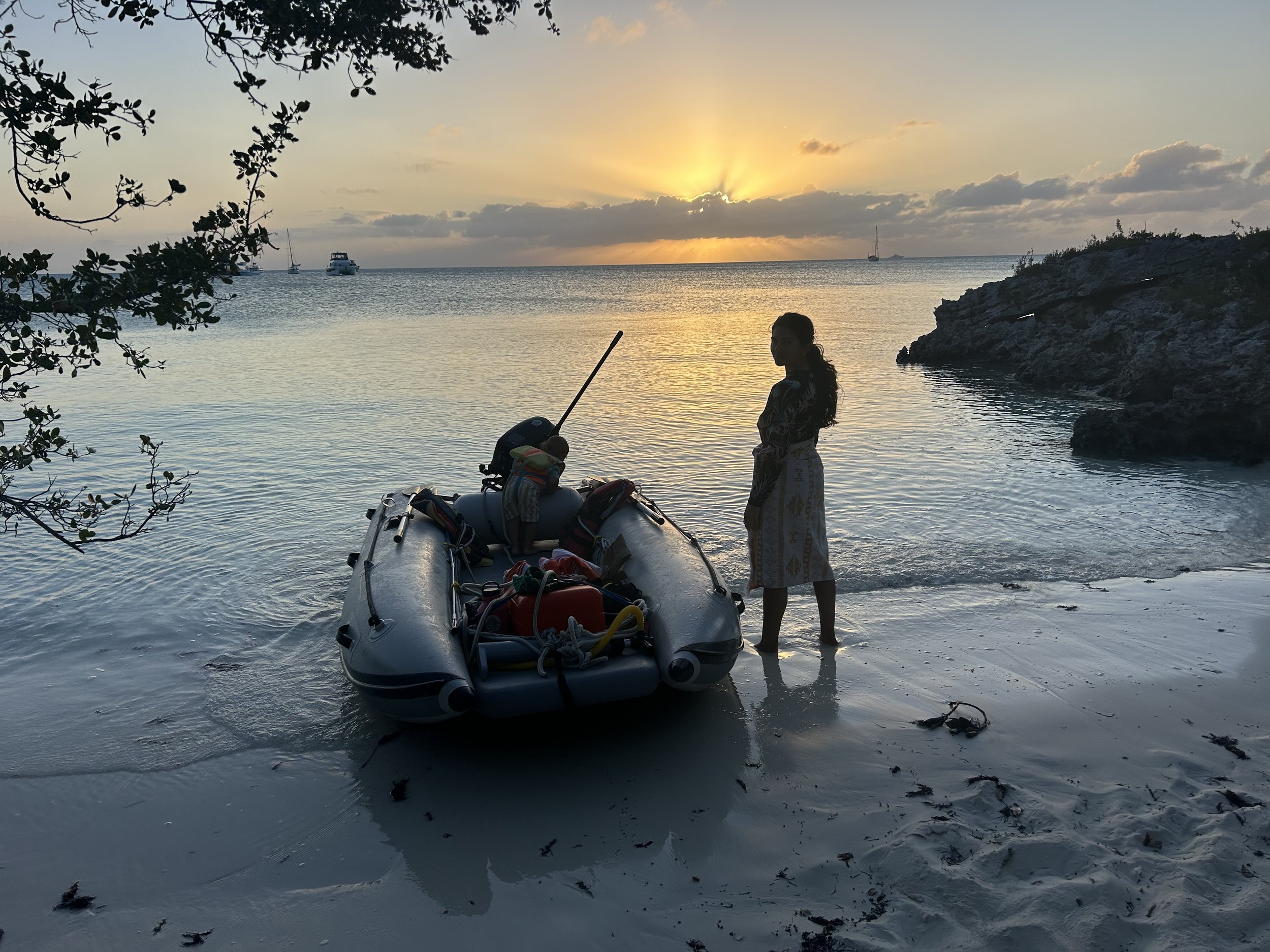 A woman stands near a small inflatable boat on a sandy shore during sunset. The sky has clouds with rays of sunlight breaking through, and there are boats in the water in the background. The scene is framed by tree branches.