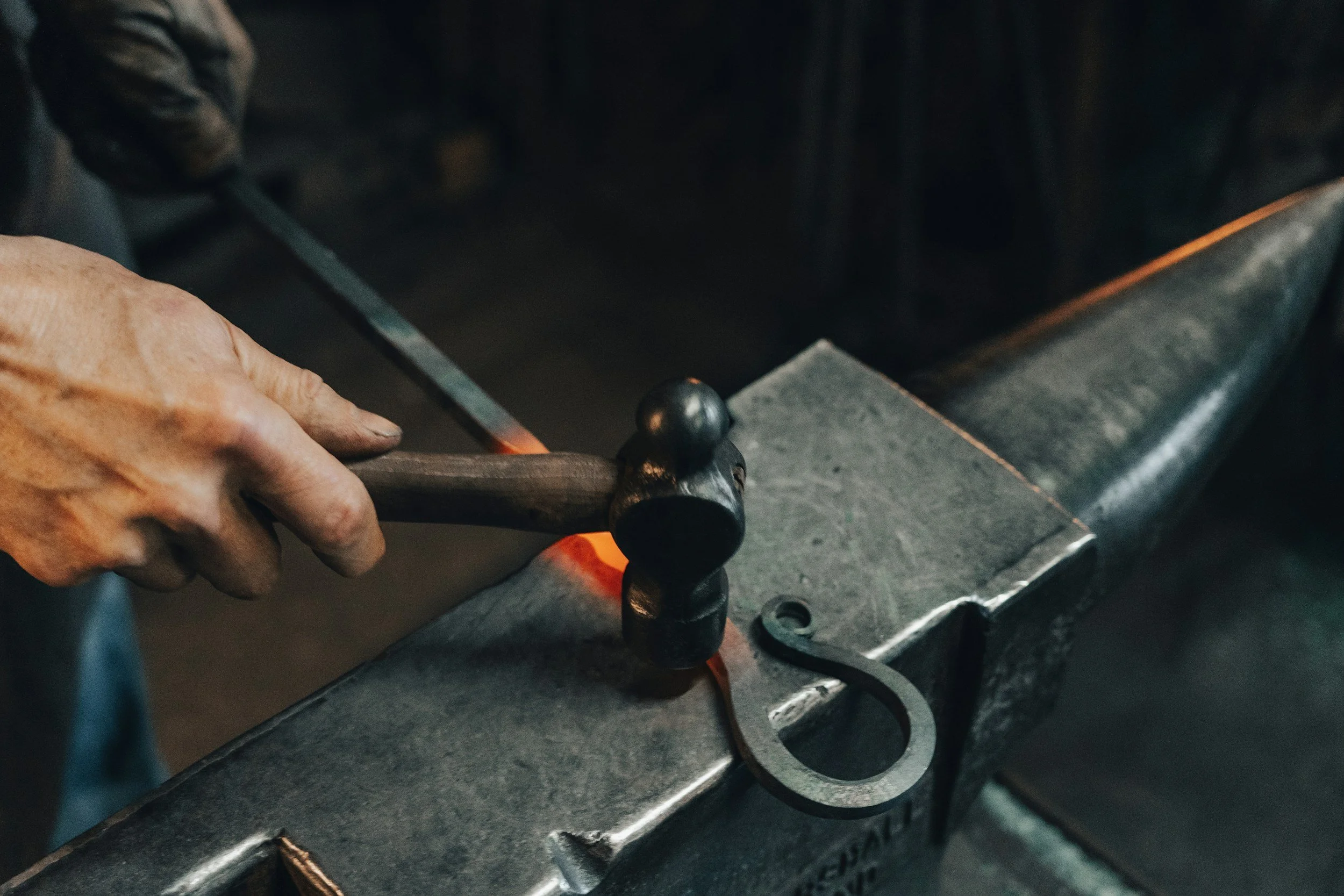 Close-up of a person's hand working with hot metal on a forging or welding bench, with tools and equipment visible in the background.