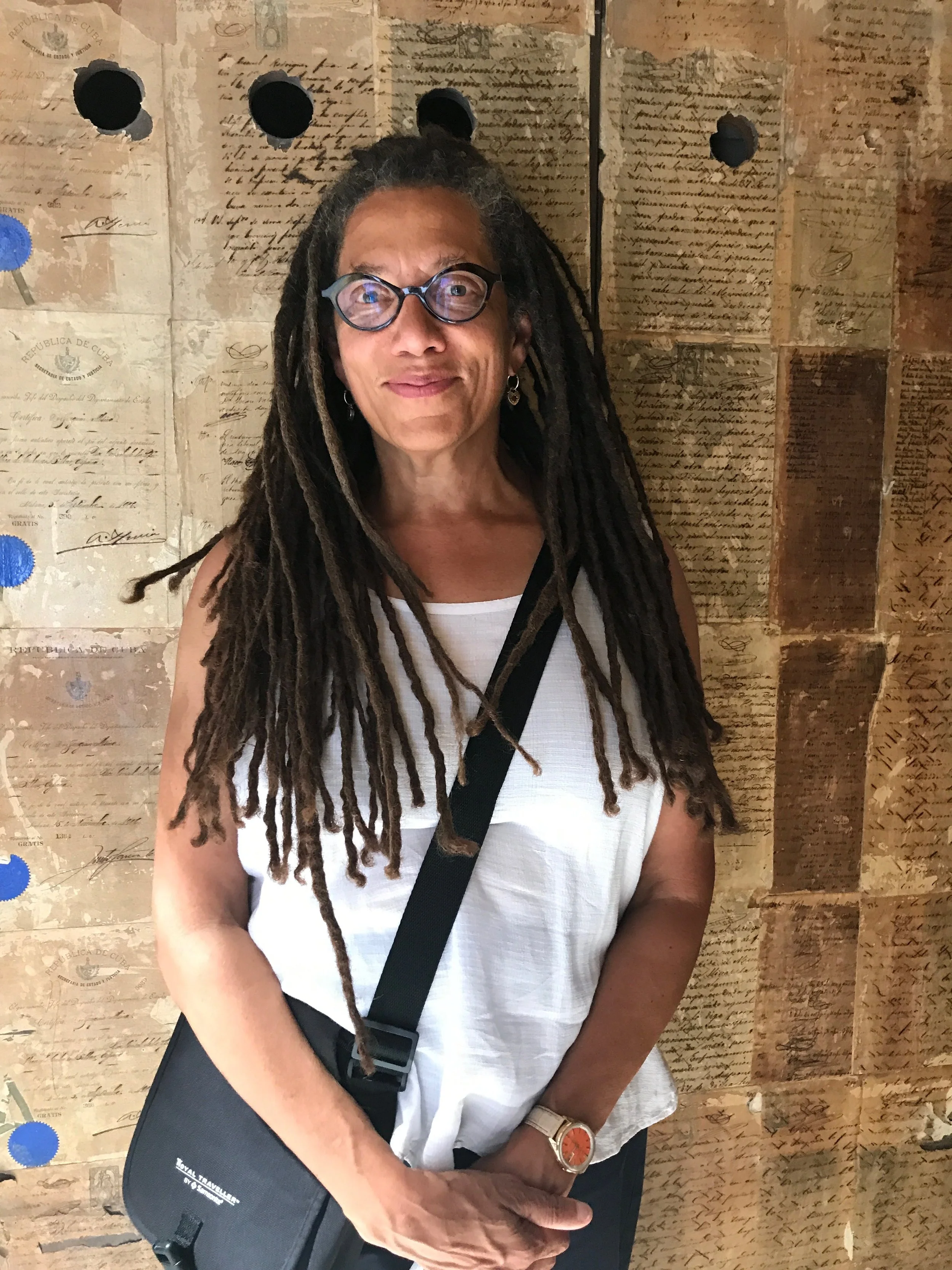 A woman with glasses and dreadlocks standing in front of a wall covered with old, handwritten letters and documents.
