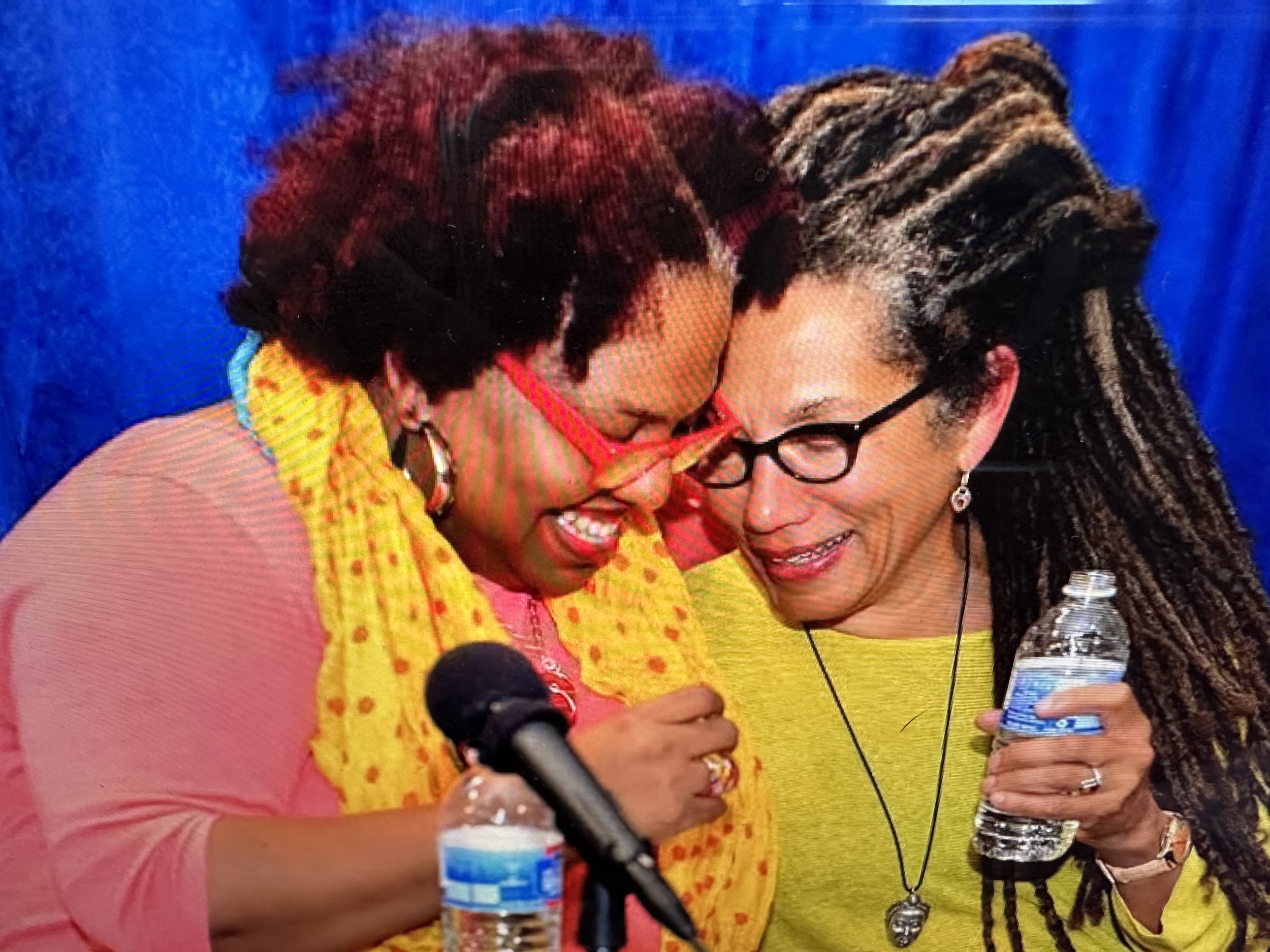 Nikky Finney and Crystal Wilkinson — two women hugging each other warmly, one holding a microphone, in a colorful setting.